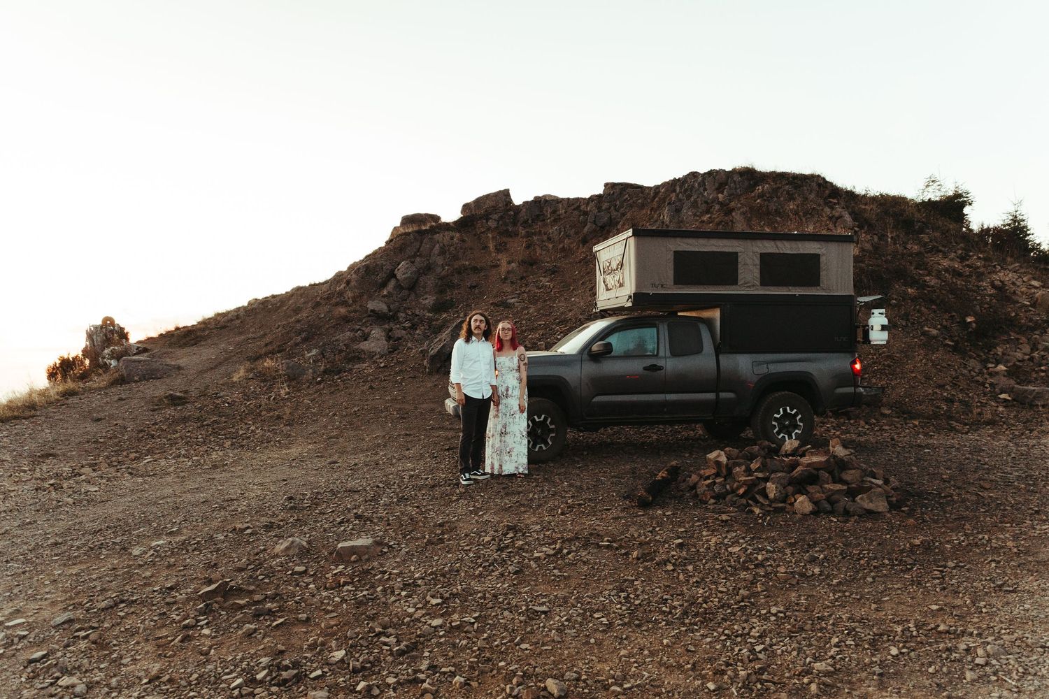 Two women stand beside a gray pickup truck with a camper shell parked on a rocky dirt hillside at sunset.