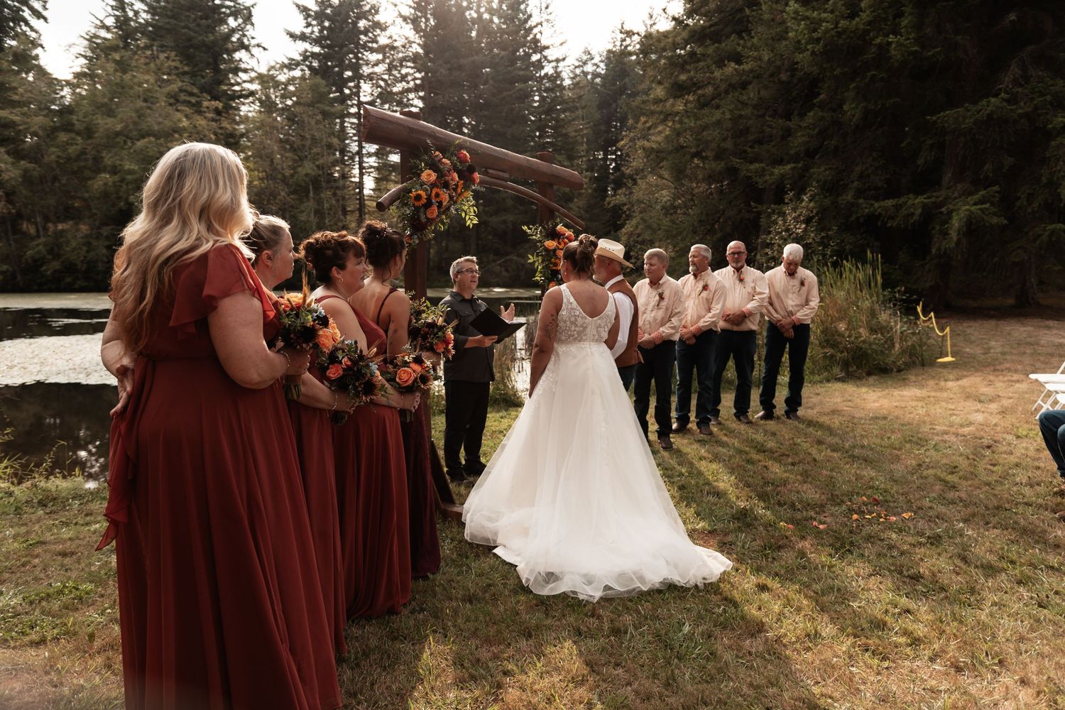 Bride walking toward groom at outdoor wedding ceremony with bridesmaids in burgundy dresses by a forest.