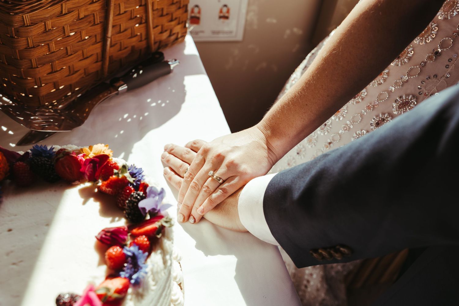 Couple holding hands with wedding rings on a sunlit white surface, surrounded by colorful floral decorations.