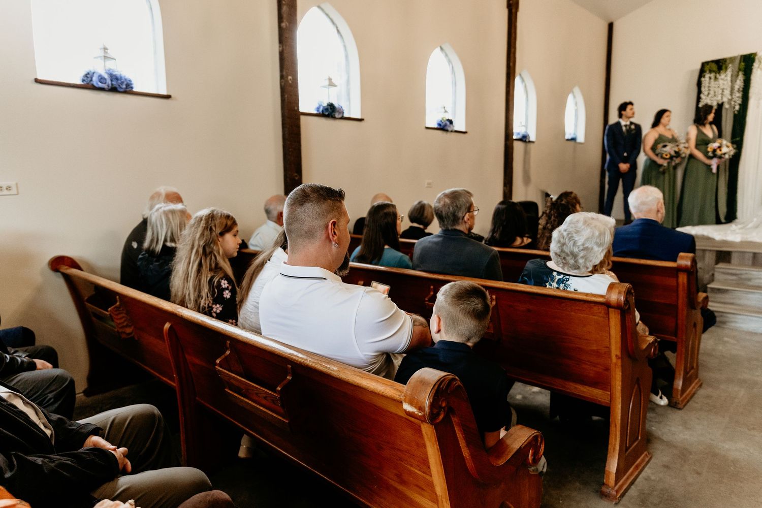 Wedding guests seated in wooden church pews watching a ceremony at a small rustic chapel altar.