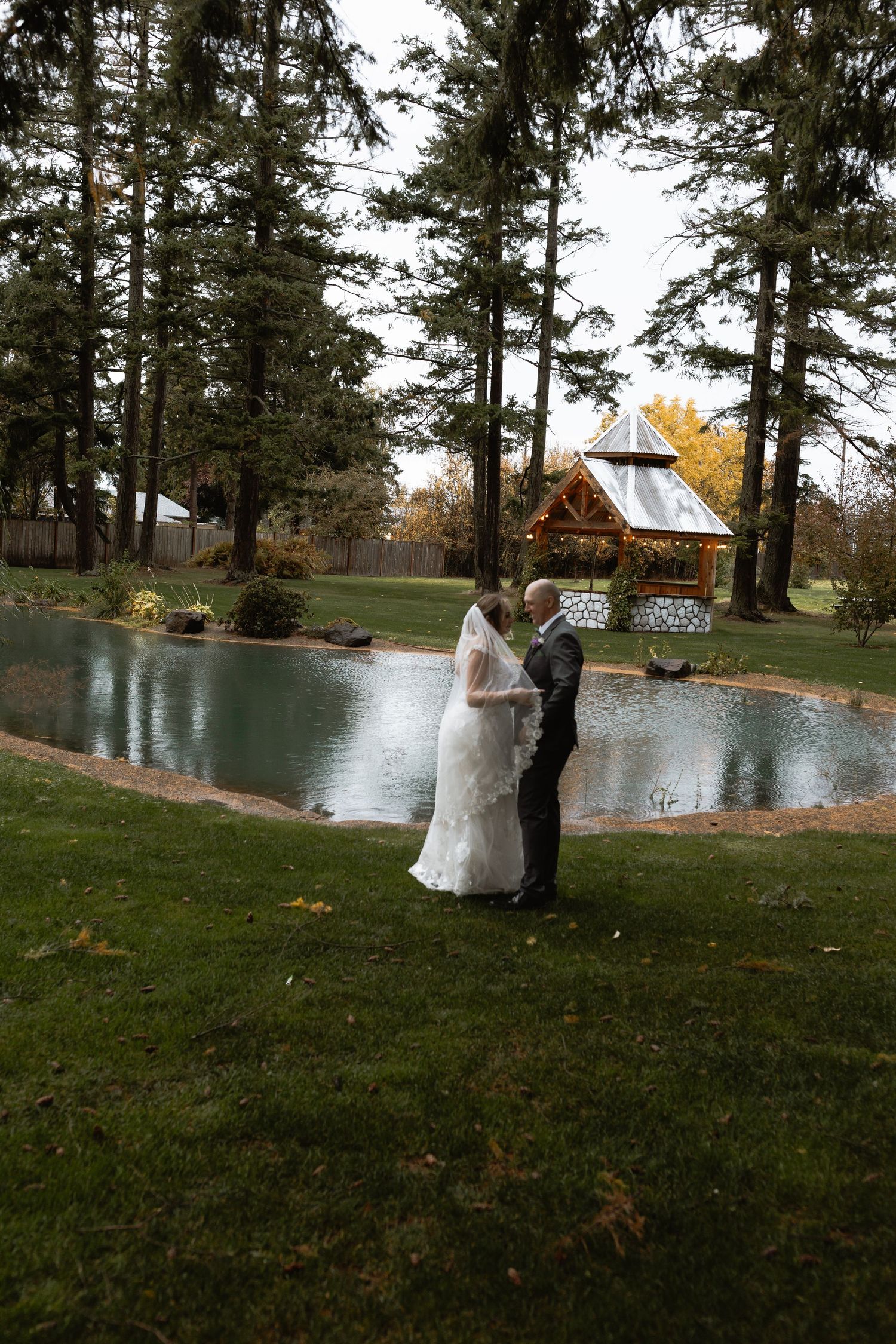 Bride and groom stand romantically by a serene pond with gazebo.