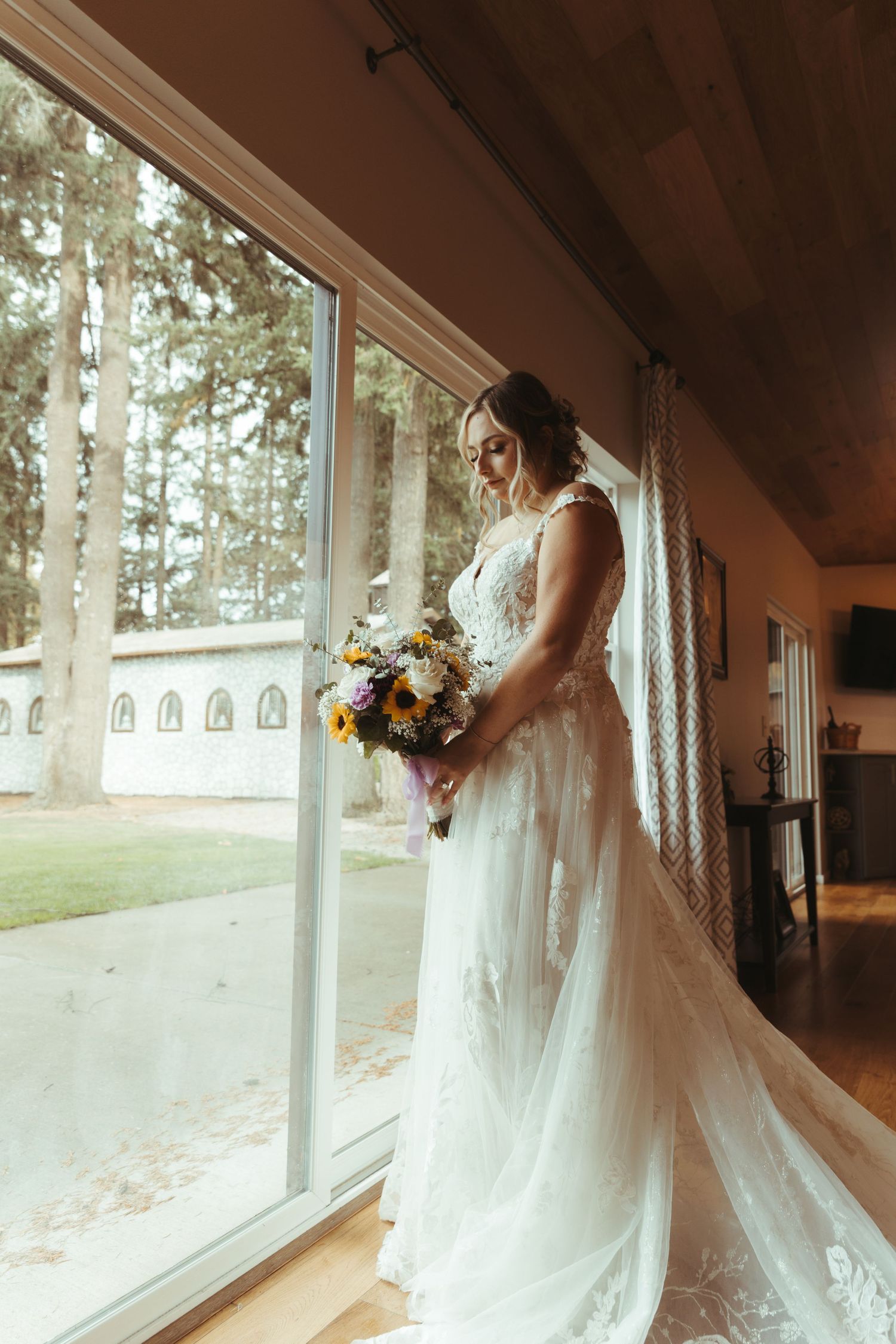 Bride in lace wedding gown holding colorful bouquet standing by large sunlit window.