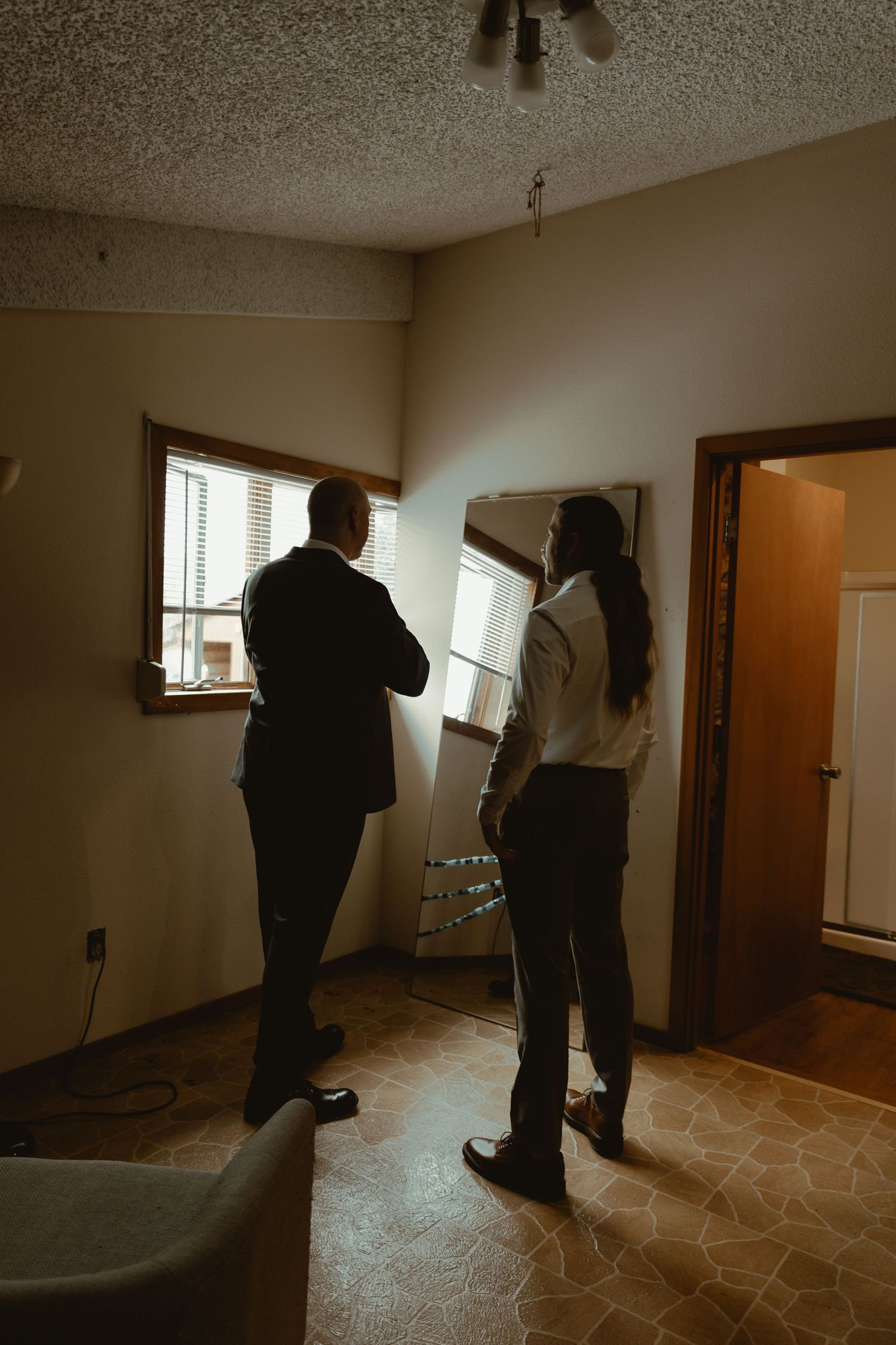 Two men in formal suits stand in silhouette near a window in a dimly lit room.