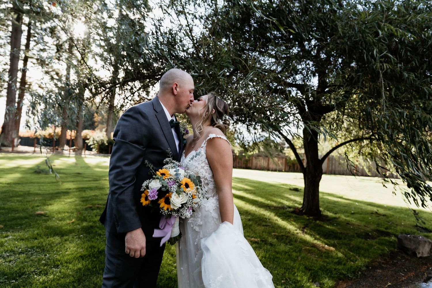 Bride and groom share romantic kiss on green lawn with colorful sunflower bouquet at outdoor wedding.