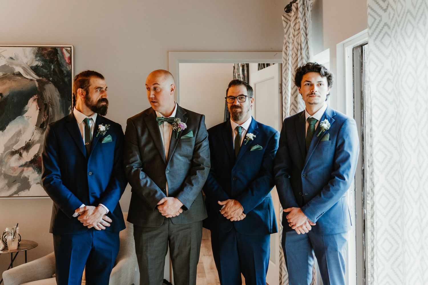 Four groomsmen in navy and gray suits with boutonnieres standing together indoors before the wedding ceremony.