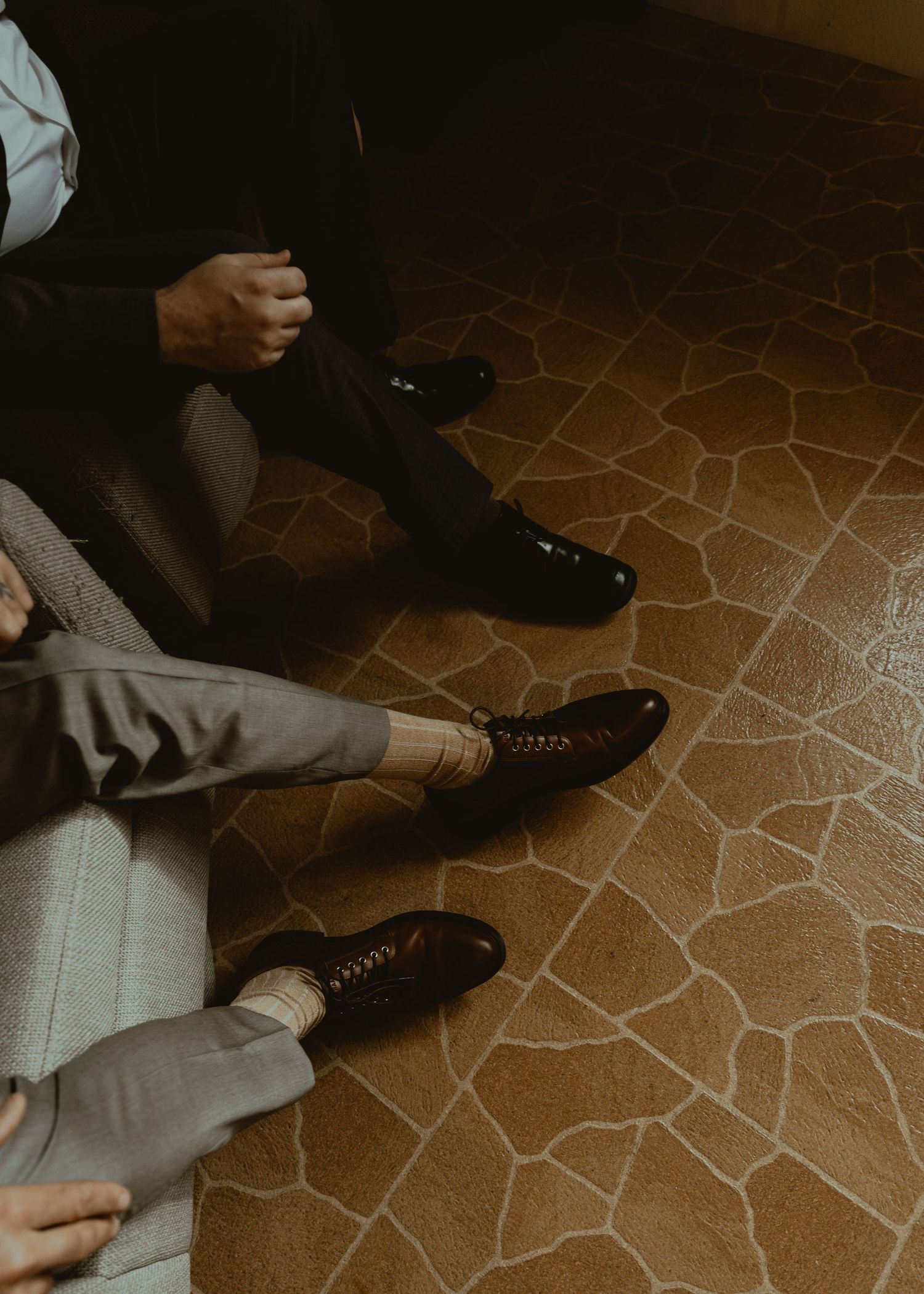 Two men in formal suits and polished oxford shoes seated on a tiled floor, showcasing elegant dress shoes.