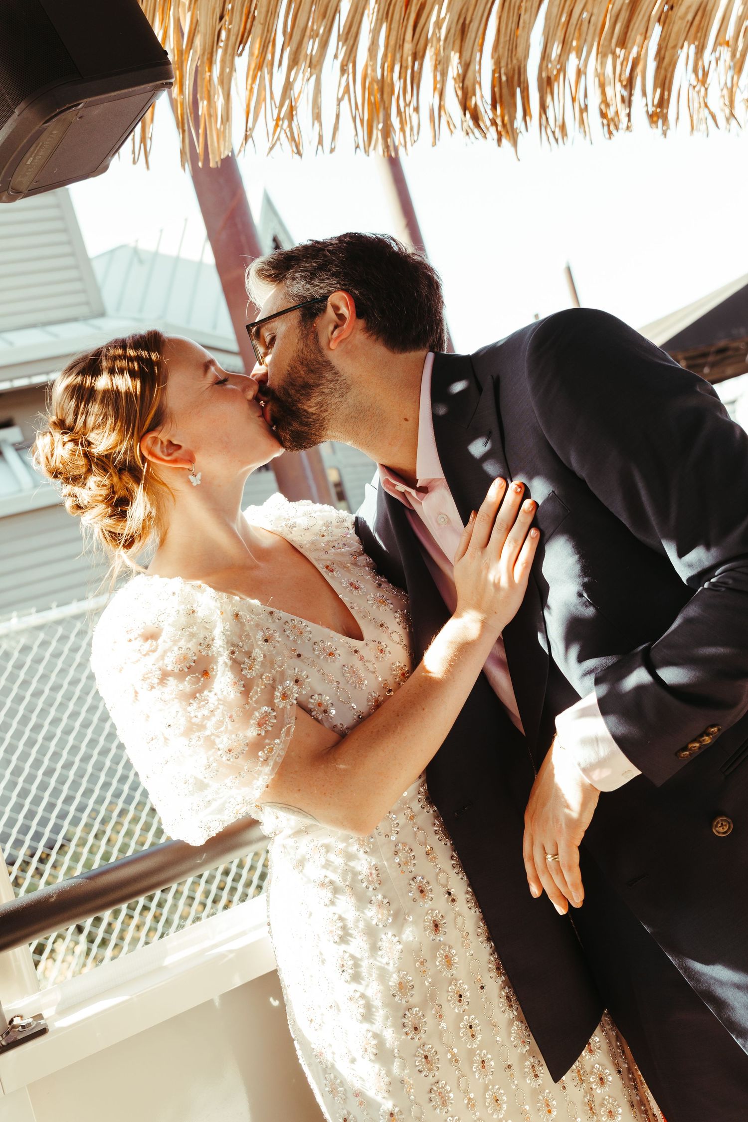 Bride and groom sharing a romantic kiss on a sunny balcony, bride in white lace dress, groom in dark suit.