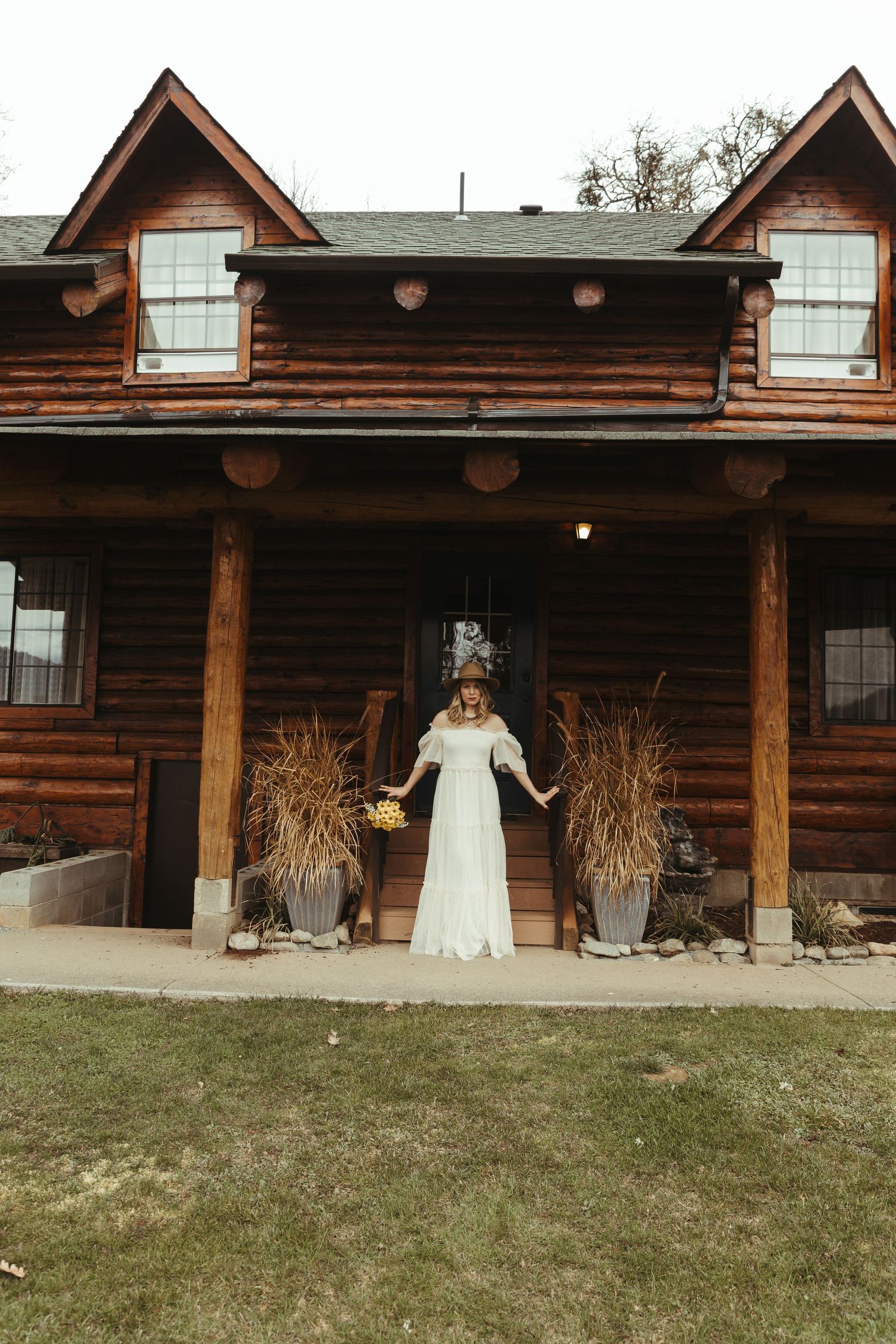 Bride in white lace gown holding bouquet stands on porch of rustic log cabin wedding venue.