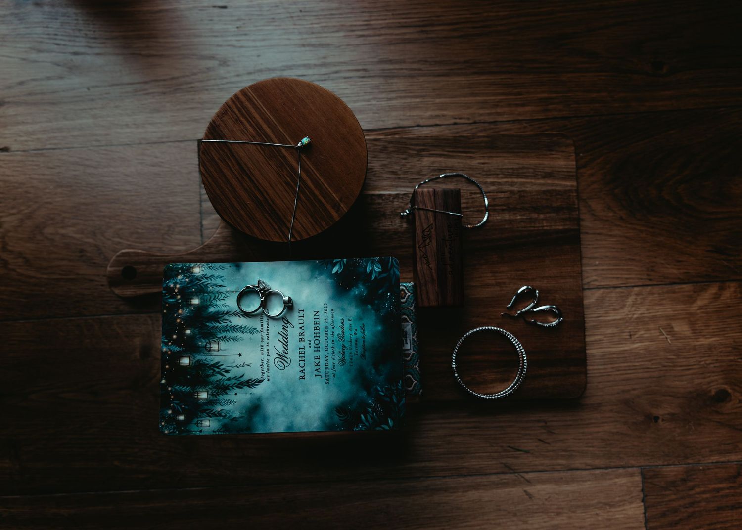 Flat lay of wedding invitation, rings, and wooden accessories on rustic hardwood floor with moody lighting.