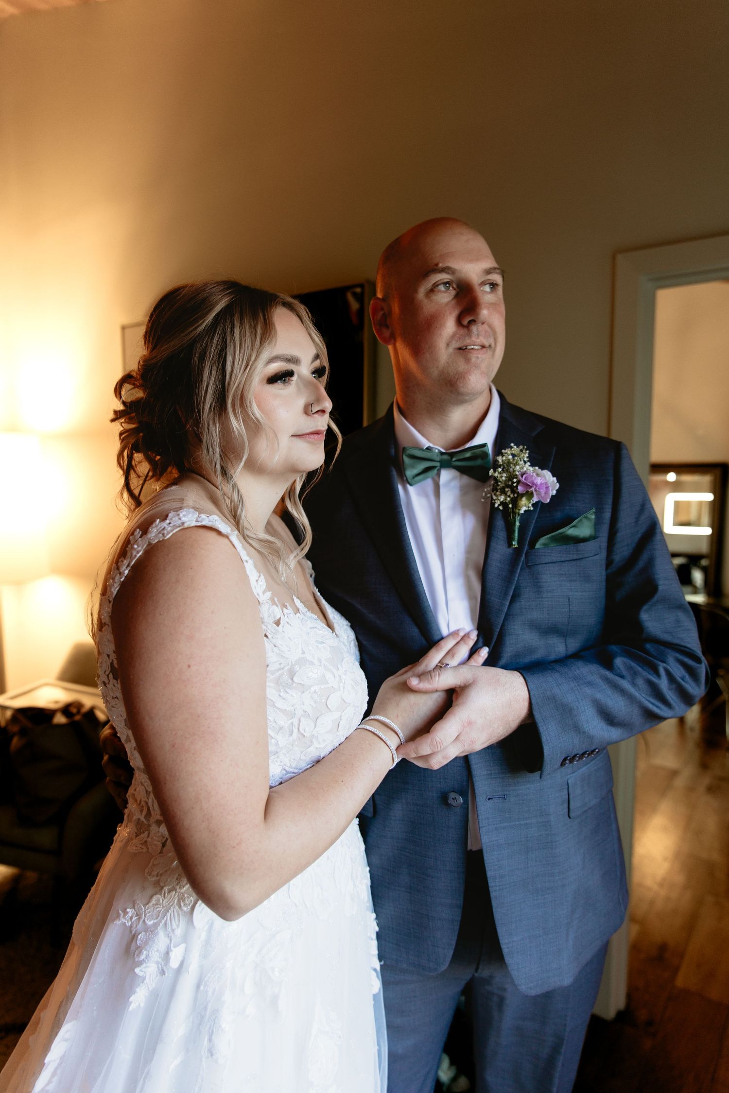 Bride in white lace gown and bald groom in blue suit holding hands indoors before wedding ceremony.