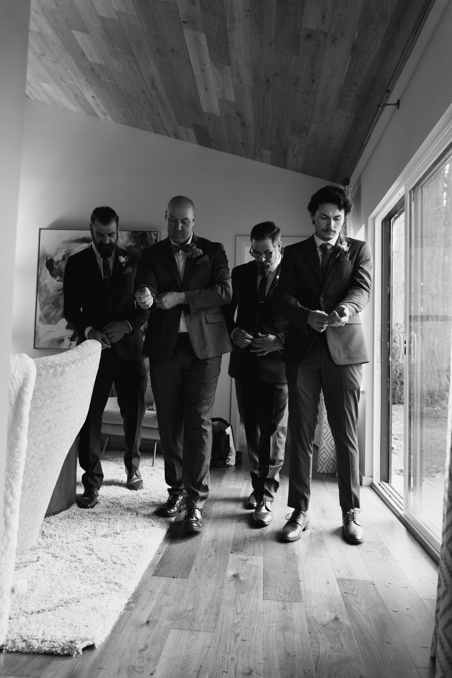 Black and white photo of four groomsmen in suits walking confidently together inside a modern venue.