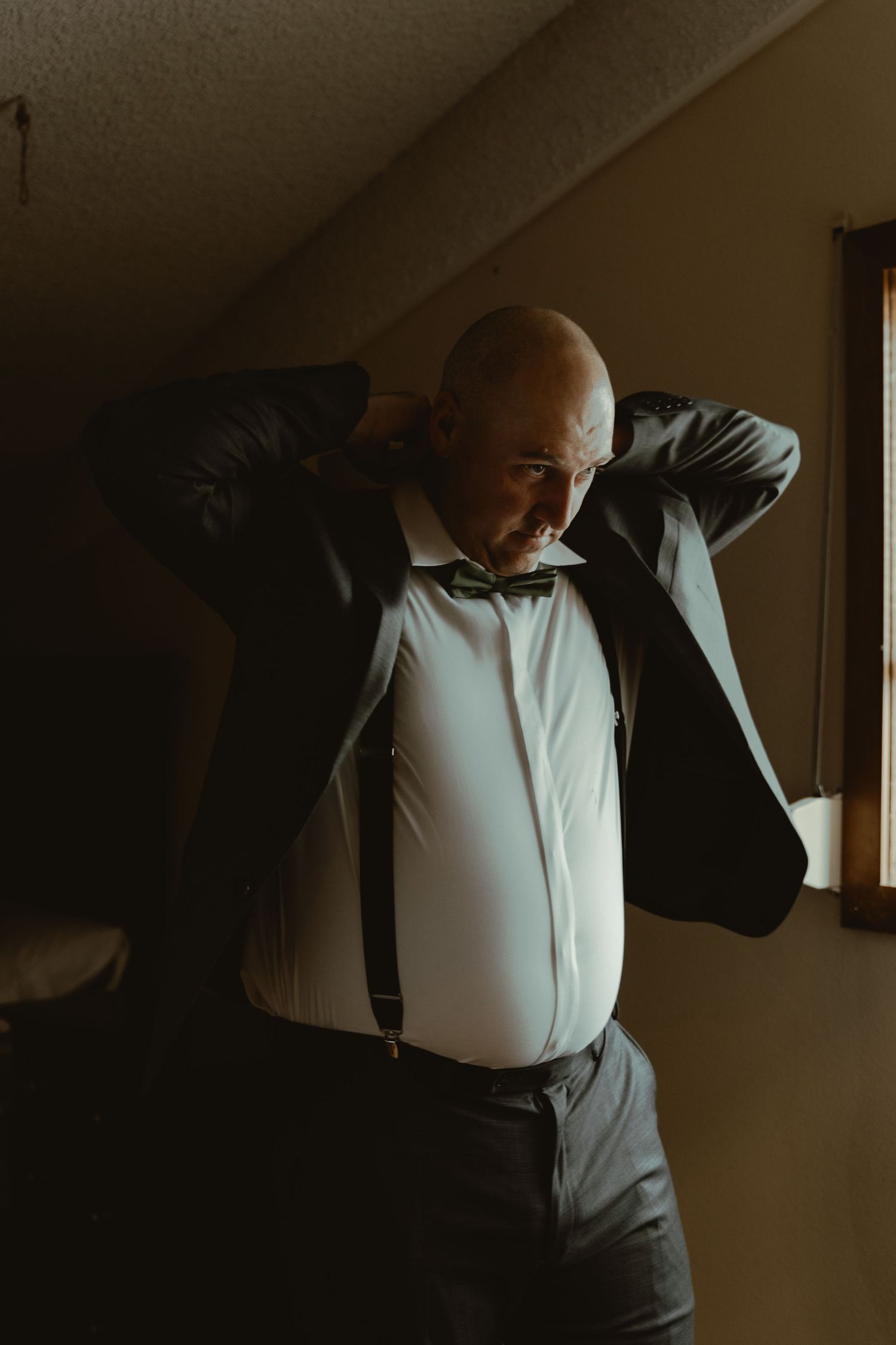 A bald groom in suspenders and bow tie puts on his suit jacket near a window.