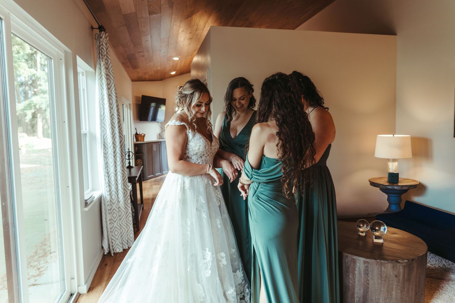 Bride smiling with three bridesmaids in green dresses gathered around her indoors.