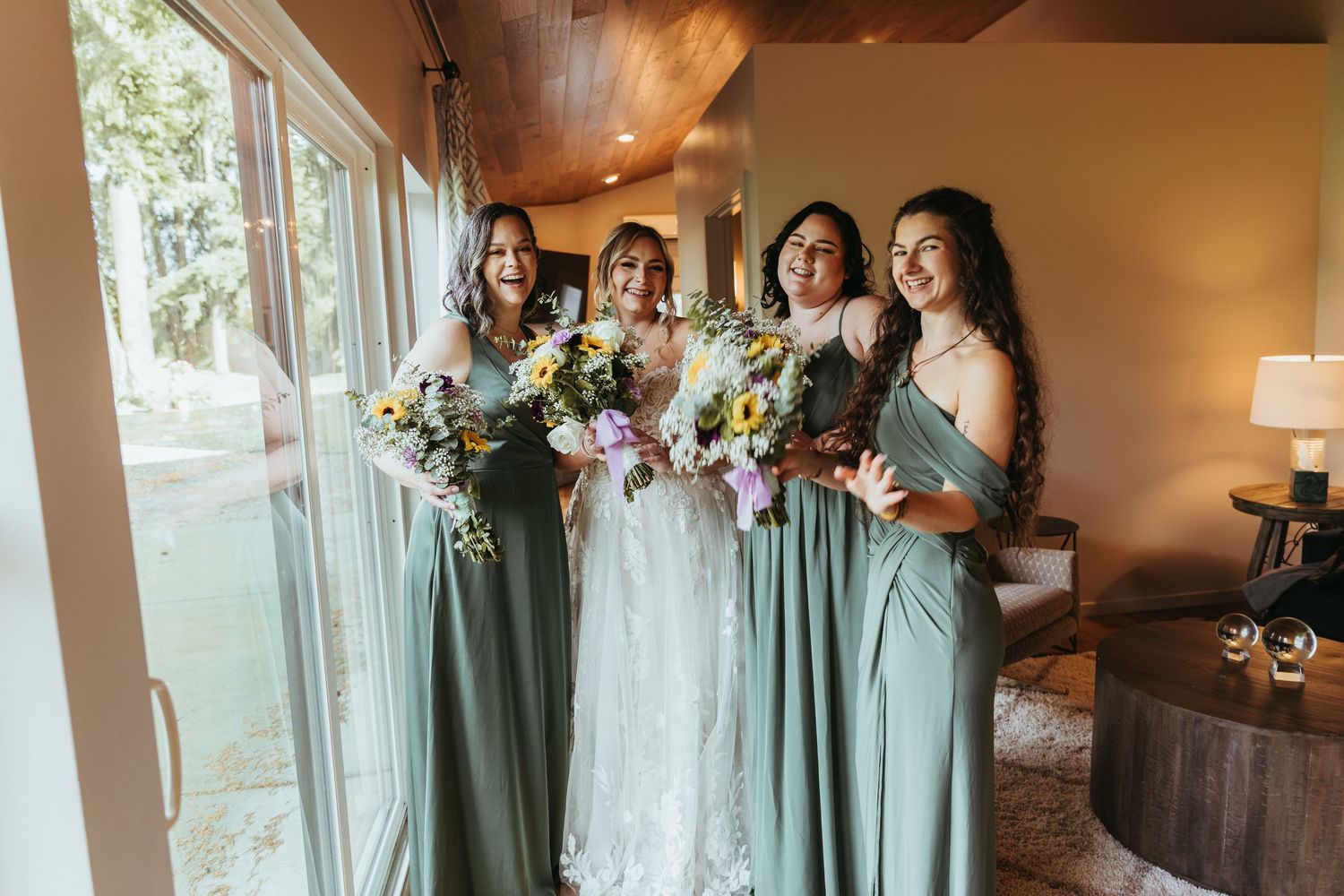 Bride and three bridesmaids in sage green gowns holding colorful bouquets near large windows in elegant venue.