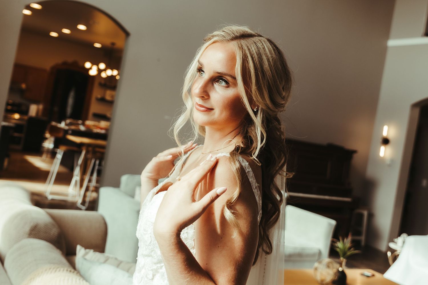 A smiling bride in a lace dress poses gracefully indoors, her wavy hair cascading over her shoulder.
