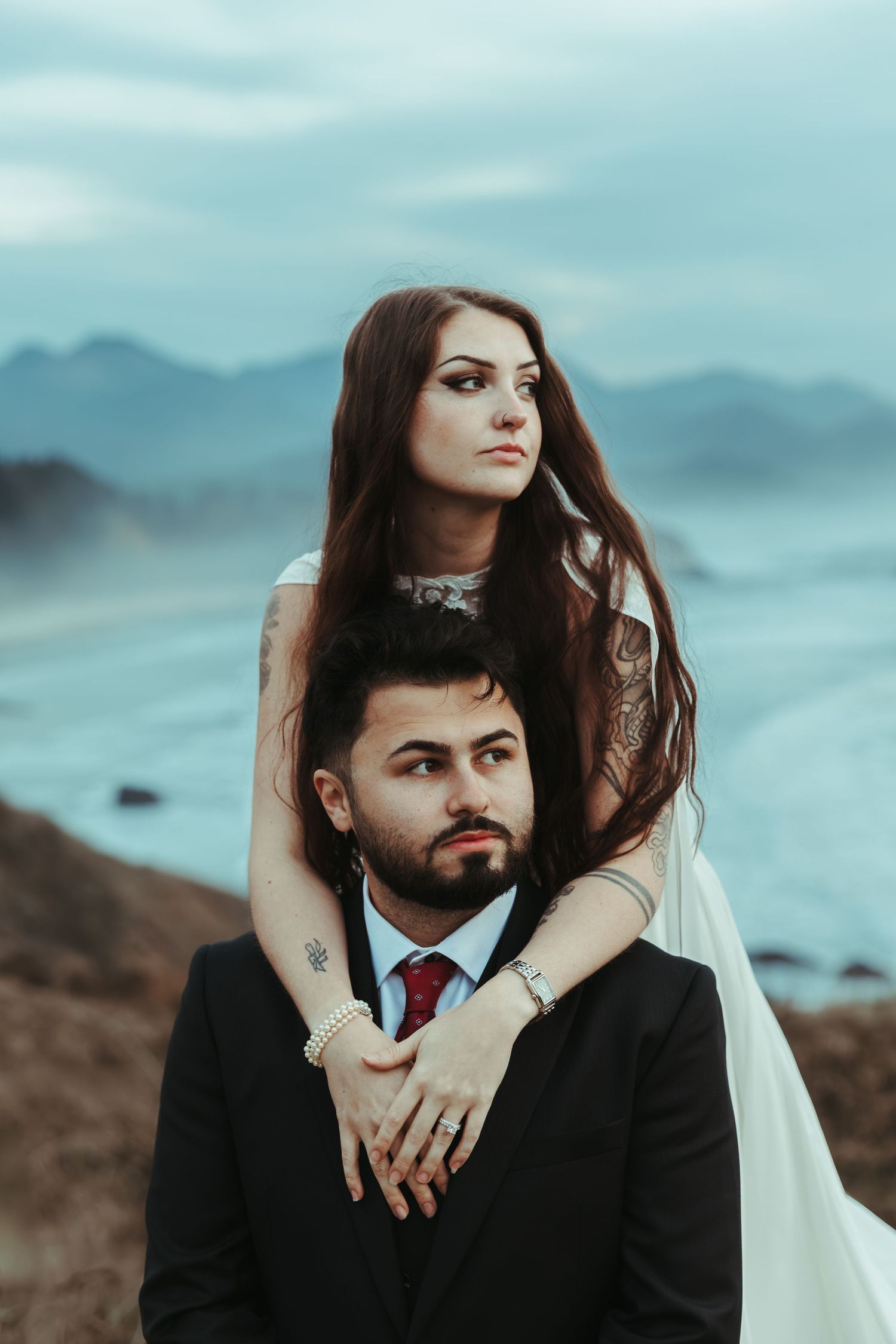 Bride in a white gown embraces a bearded groom in a black suit and red tie from behind on a misty coastal cliff overlooking the ocean.