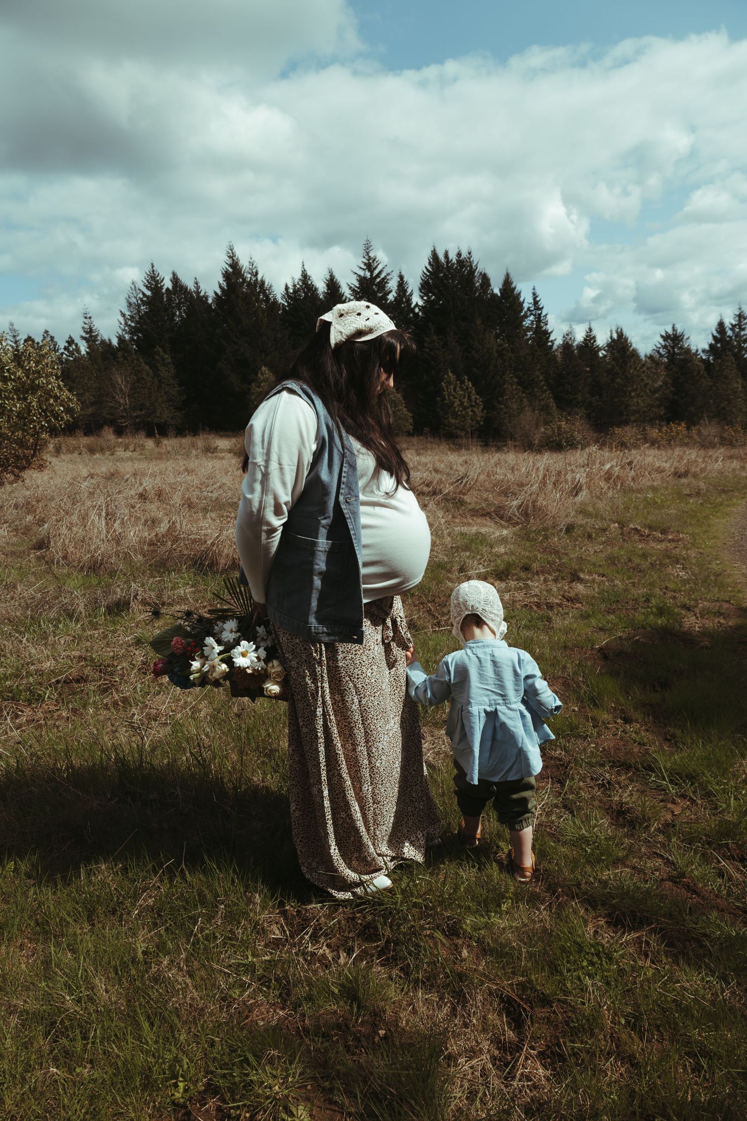 Pregnant woman holding flowers walks with toddler in meadow near pine forest on cloudy day.
