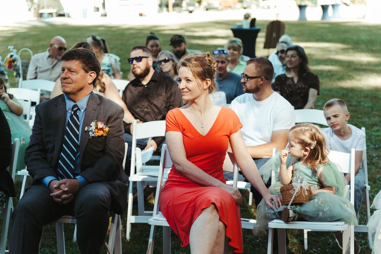 Guests seated in white chairs at an outdoor wedding ceremony, woman in red dress and man in suit in foreground.