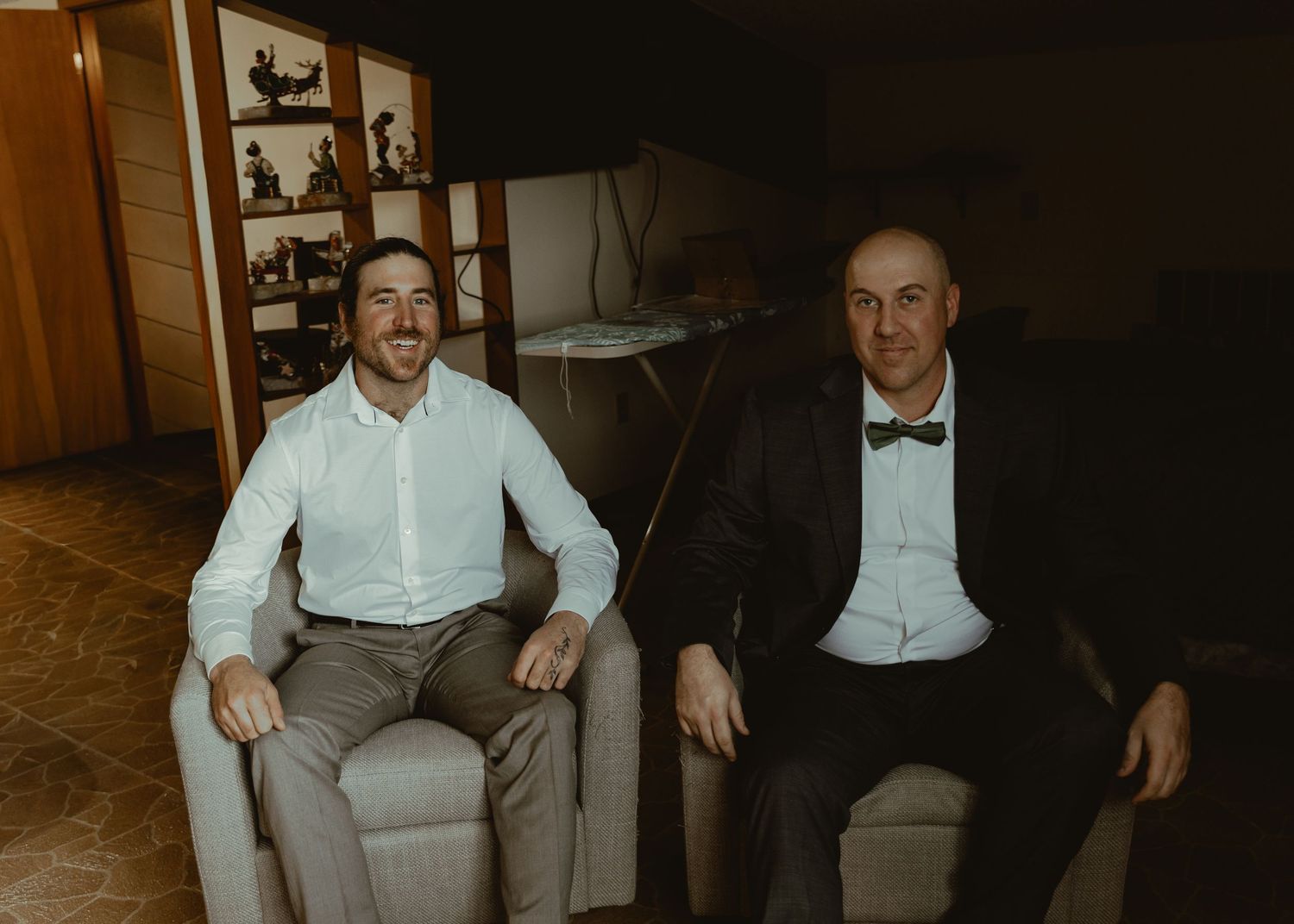Two men in formal attire smile while seated in chairs inside an elegant room.
