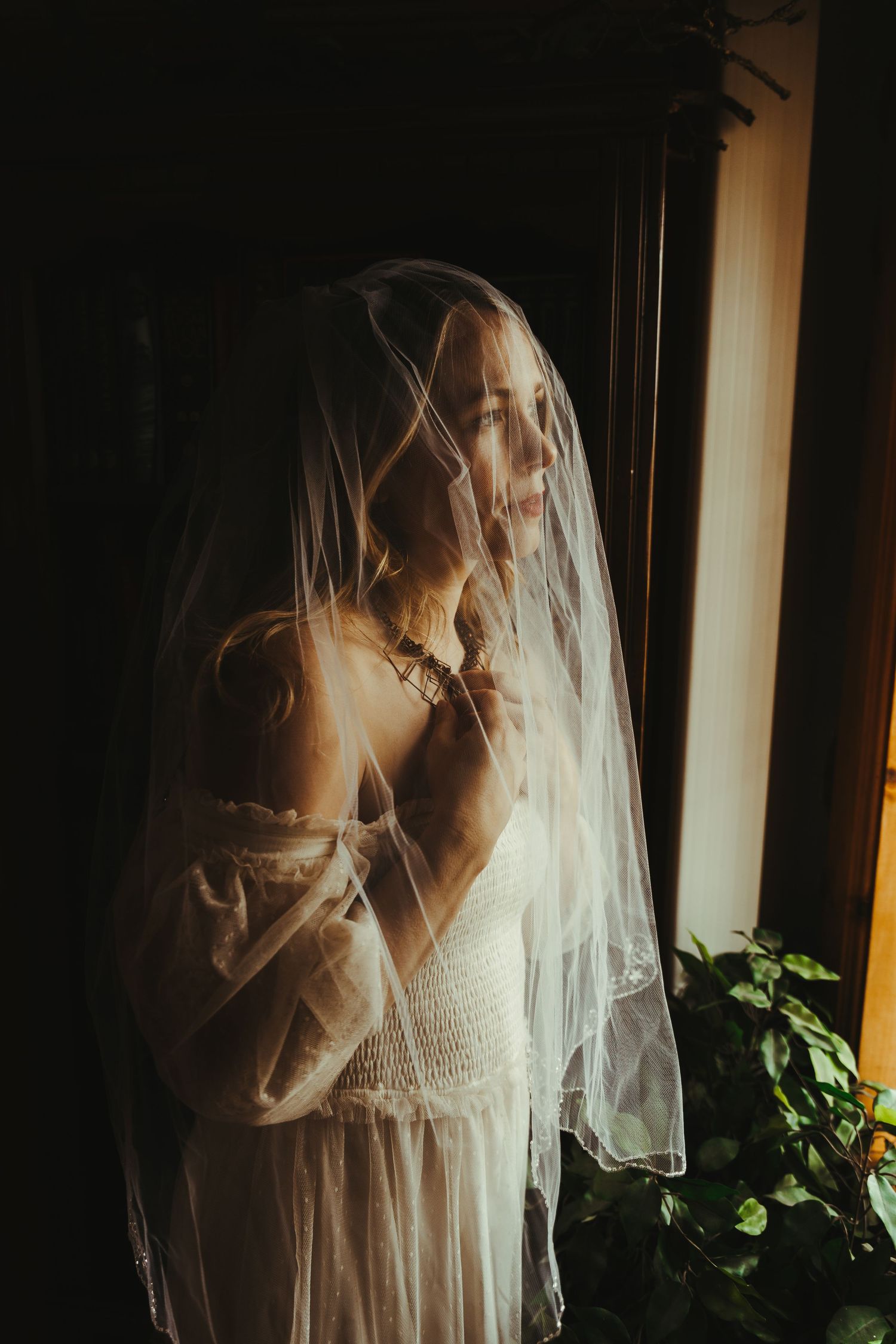 A bride in a white veil and dress stands by a window in soft golden light, gazing thoughtfully to the side.