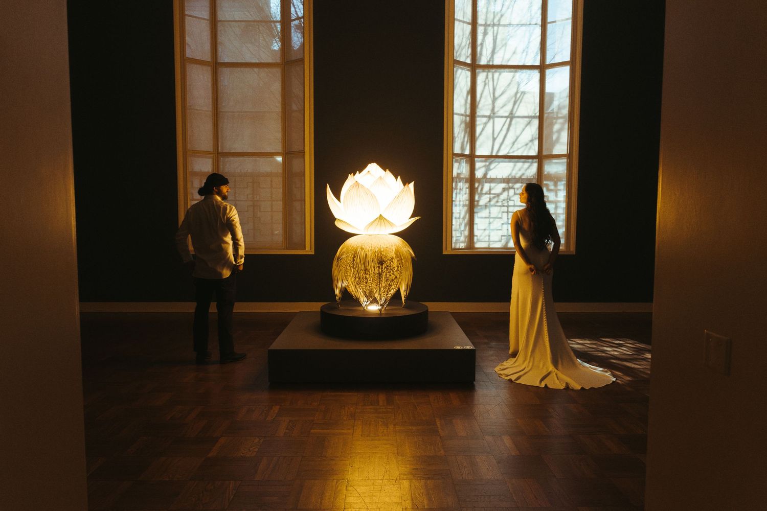 Couple standing beside a glowing illuminated lotus flower sculpture in a dimly lit gallery.
