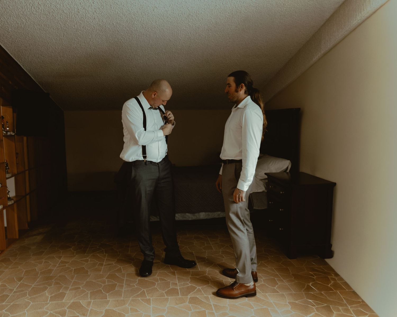 Two men in formal wedding attire converse in a dimly lit hotel room while getting ready.
