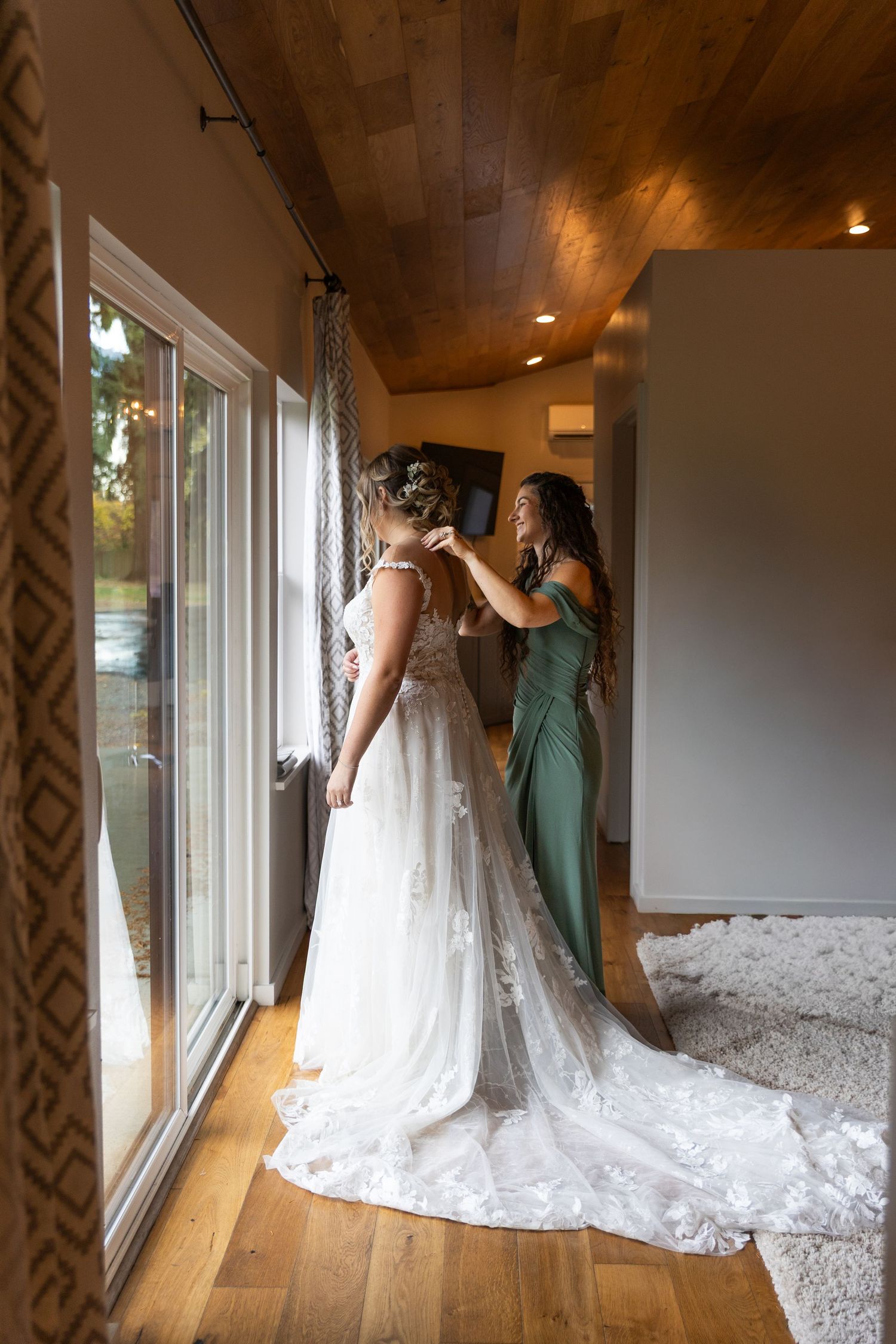 Bridesmaid in green dress fastening back of bride's white lace gown by large window.