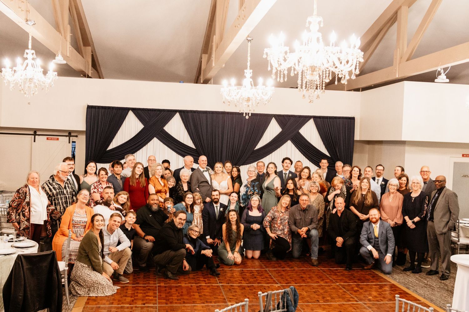 Large wedding group photo with guests posing together in an elegant venue with chandeliers and dark draping.