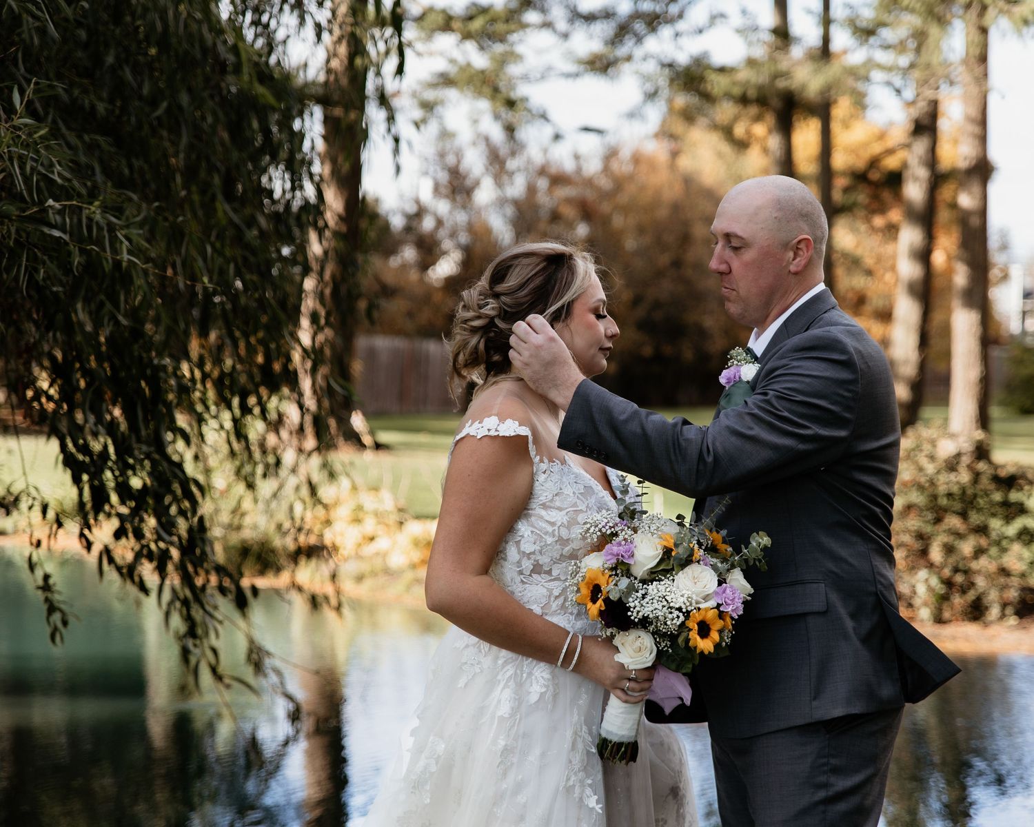 Groom tenderly touches bride's face near a pond surrounded by autumn foliage at outdoor wedding.