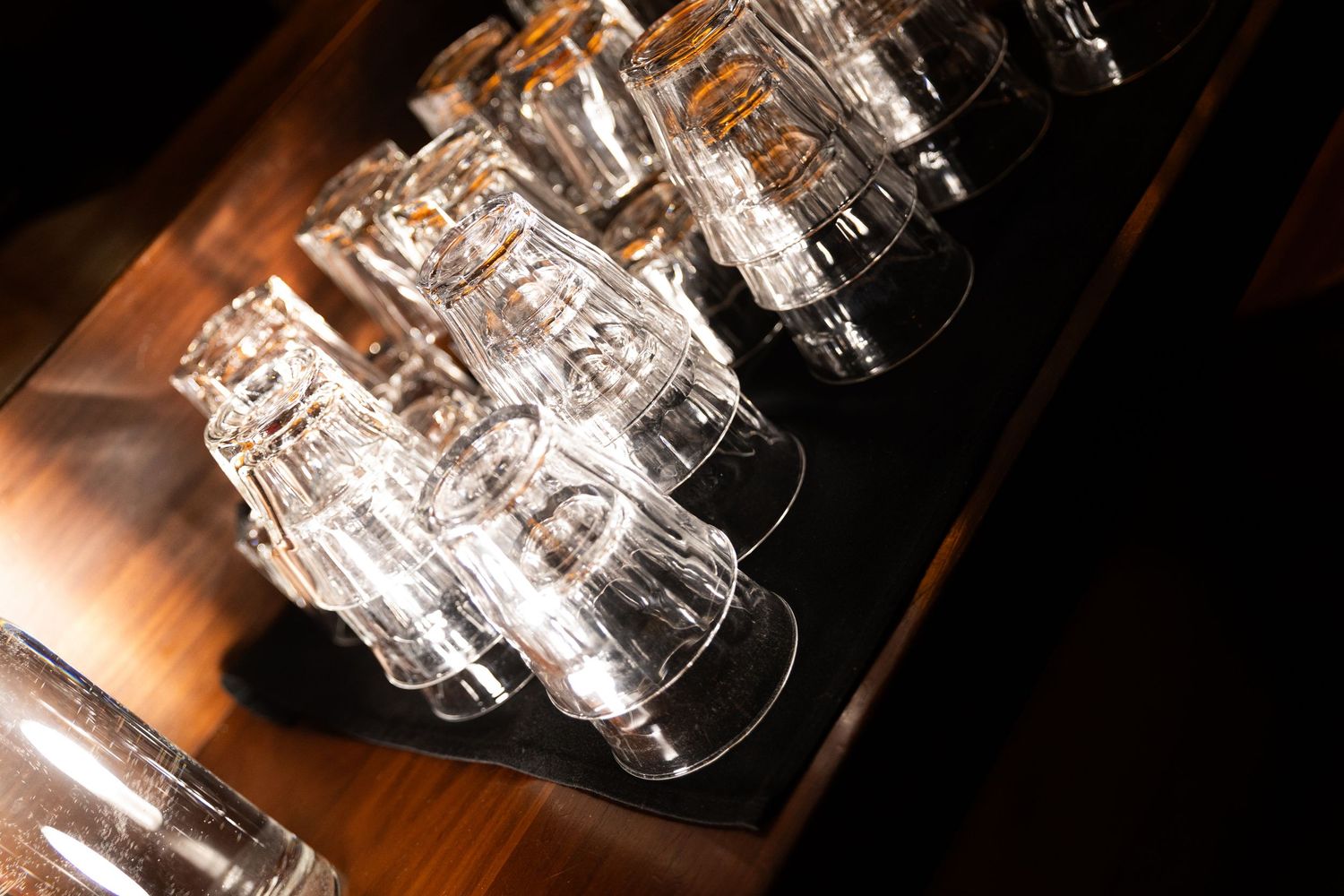 Clear glass tumblers stacked upside down on a dark wooden bar surface, illuminated by warm ambient lighting.