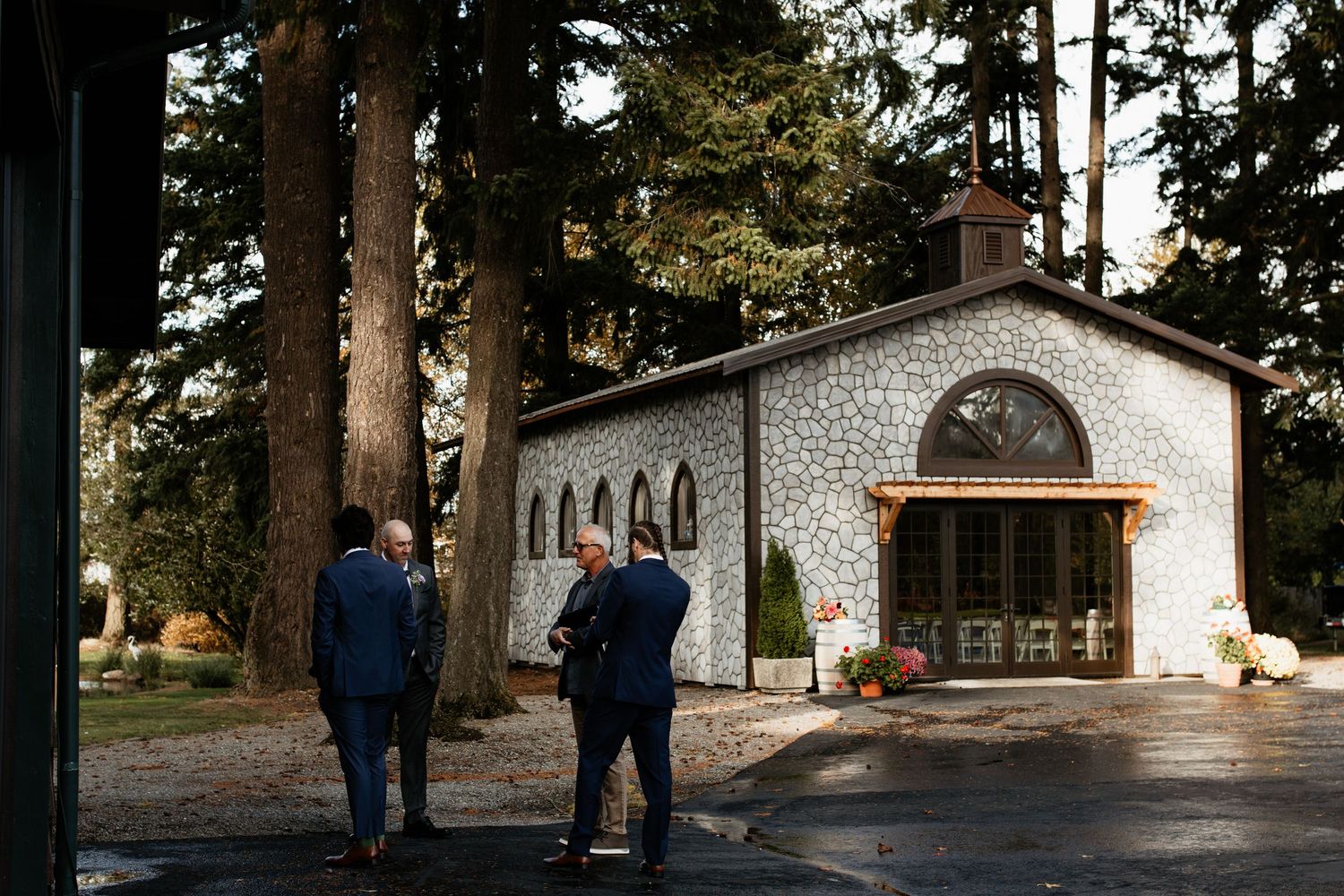 Guests gather outside a charming stone chapel surrounded by tall trees at golden hour.