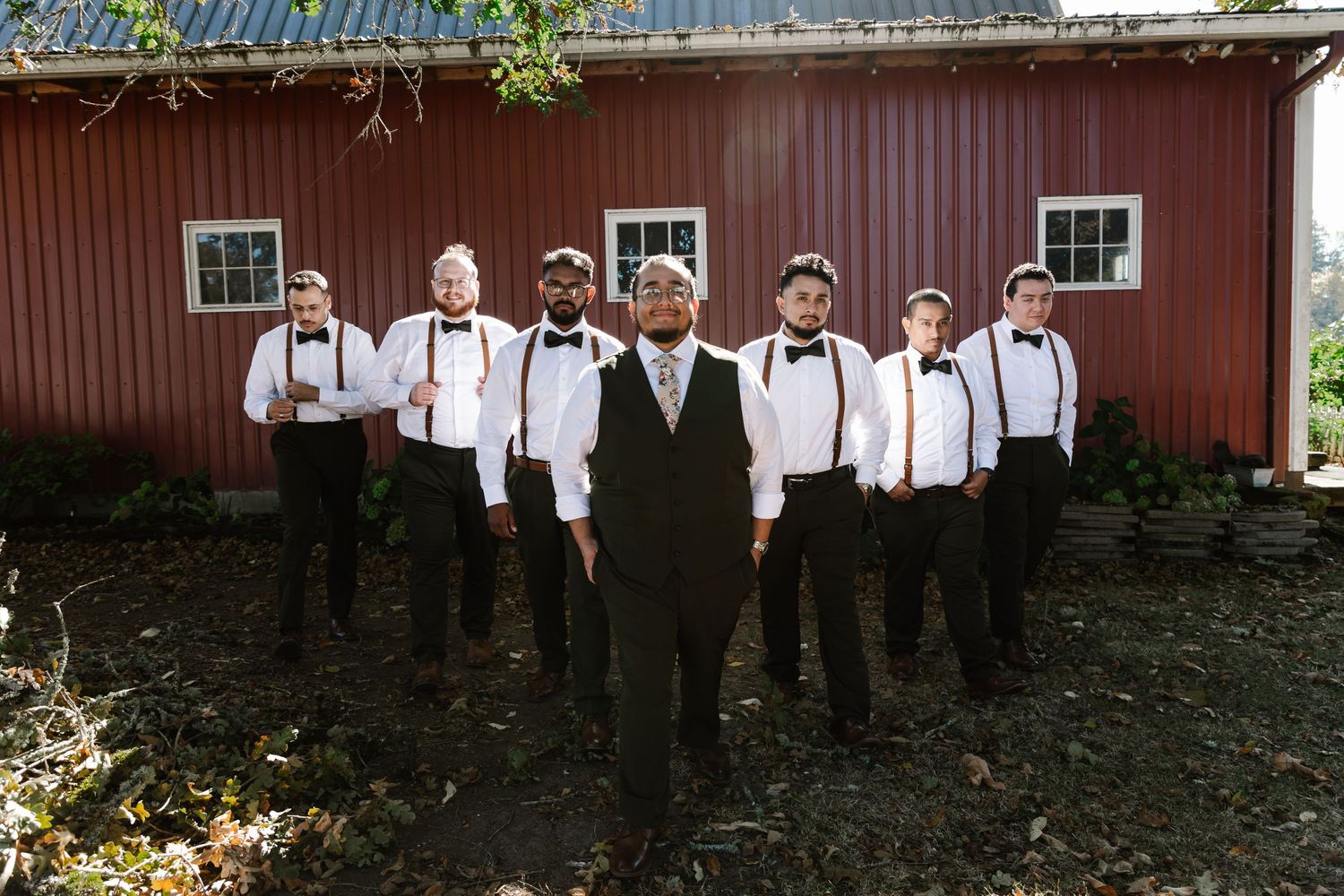 Seven groomsmen in white shirts, black vests and bow ties stand together in front of a red barn on a sunny day.