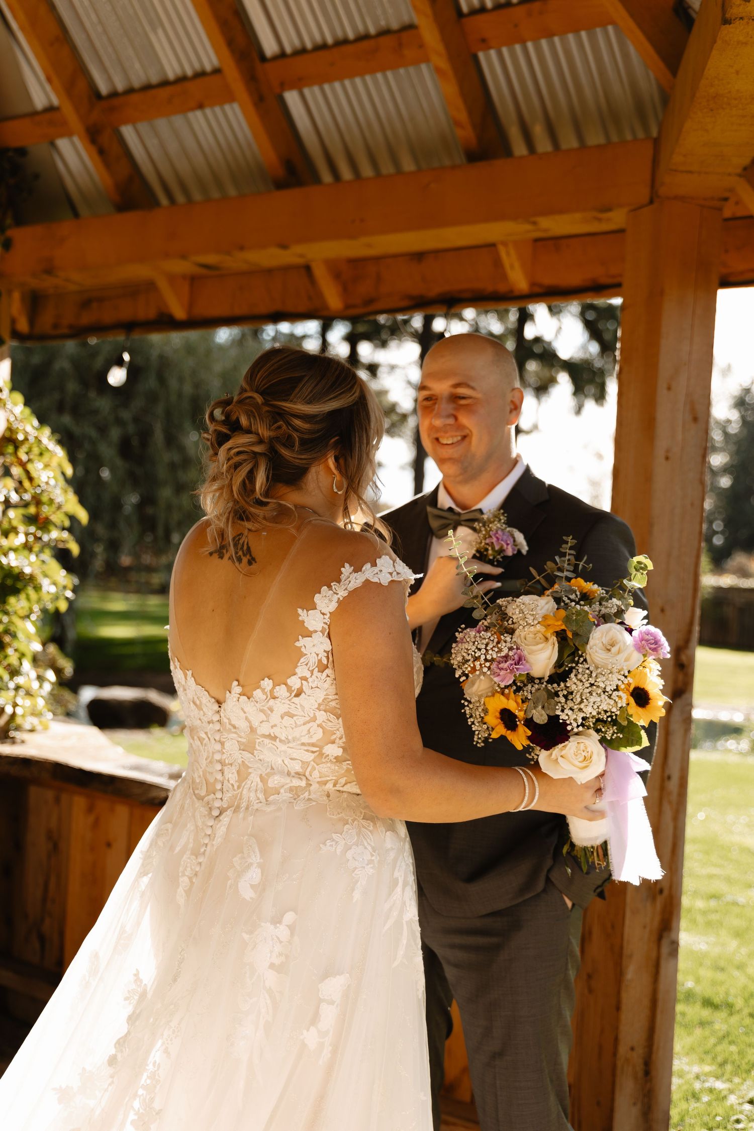 Groom smiles joyfully as bride in lace wedding gown holds colorful sunflower bouquet under wooden gazebo.