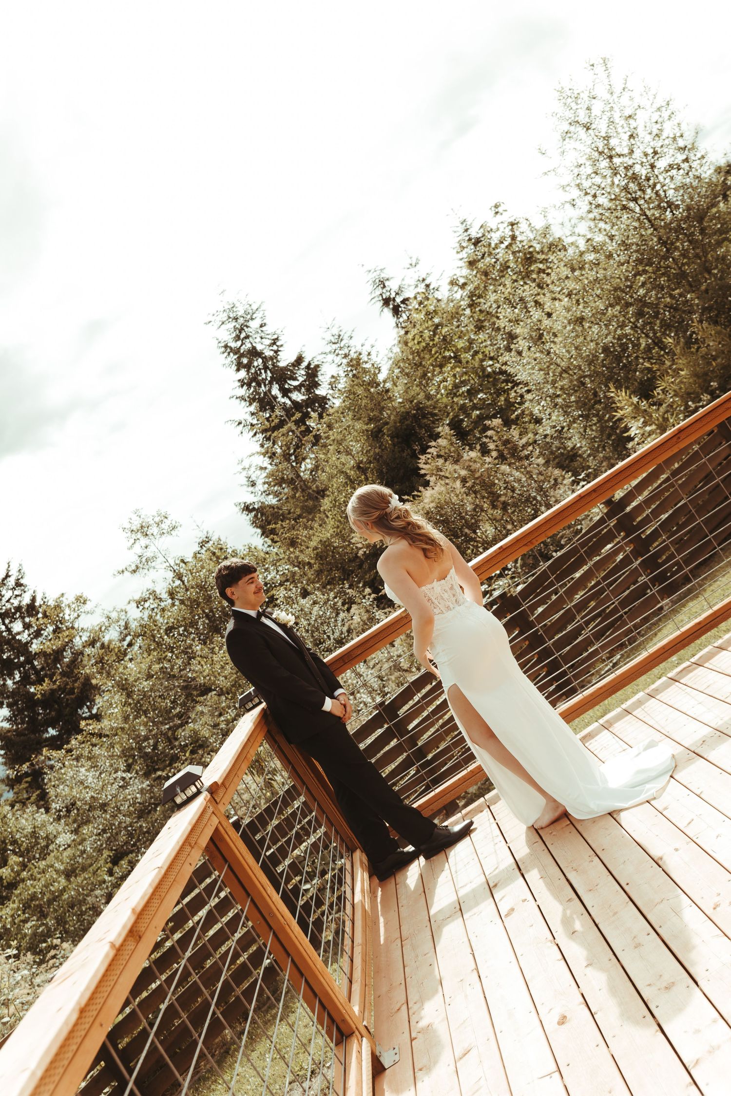 Bride and groom standing on a wooden deck surrounded by lush green trees on their wedding day.