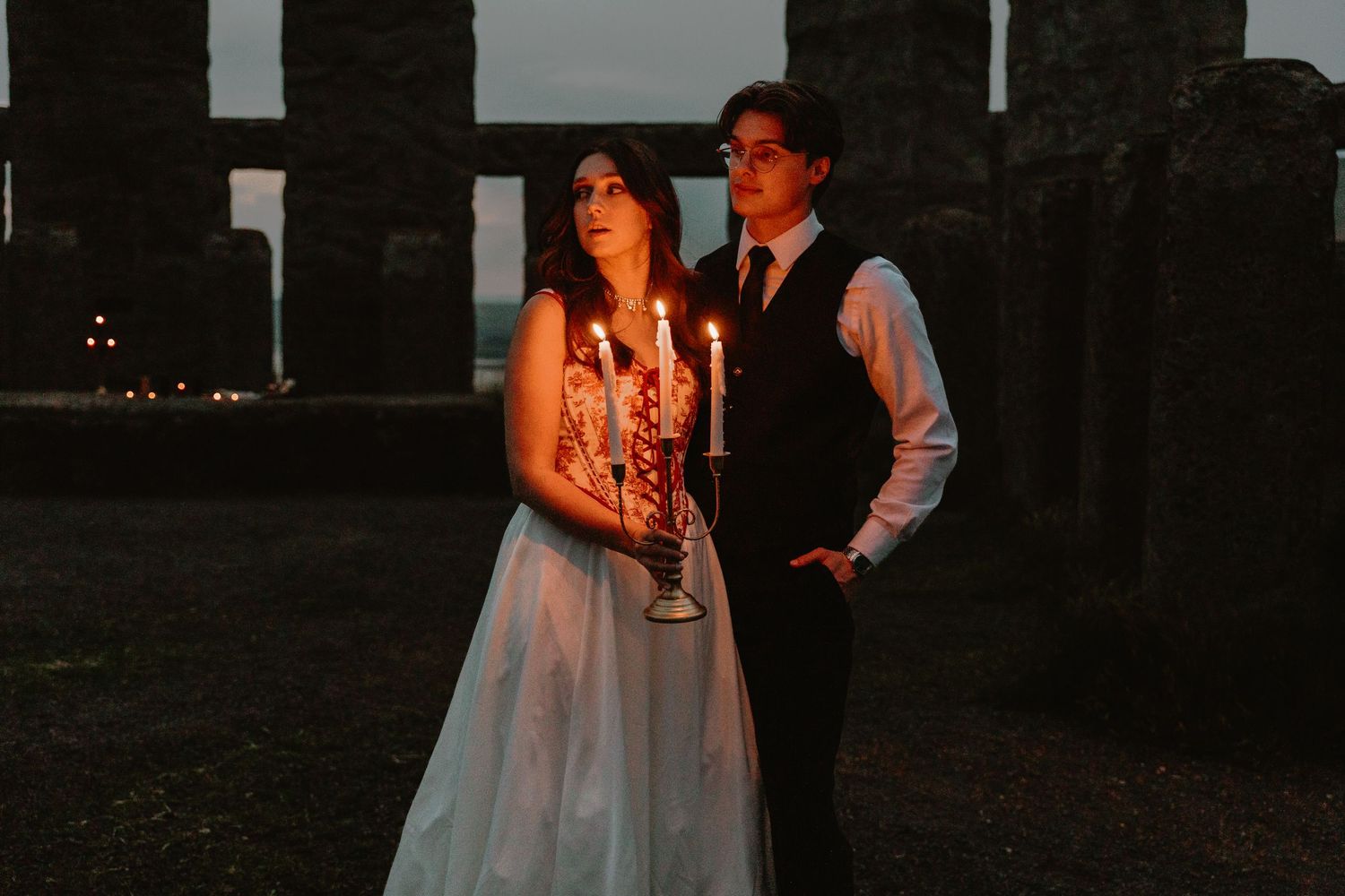 Couple in wedding attire holds a candelabra with lit candles at dusk near ancient stone ruins.