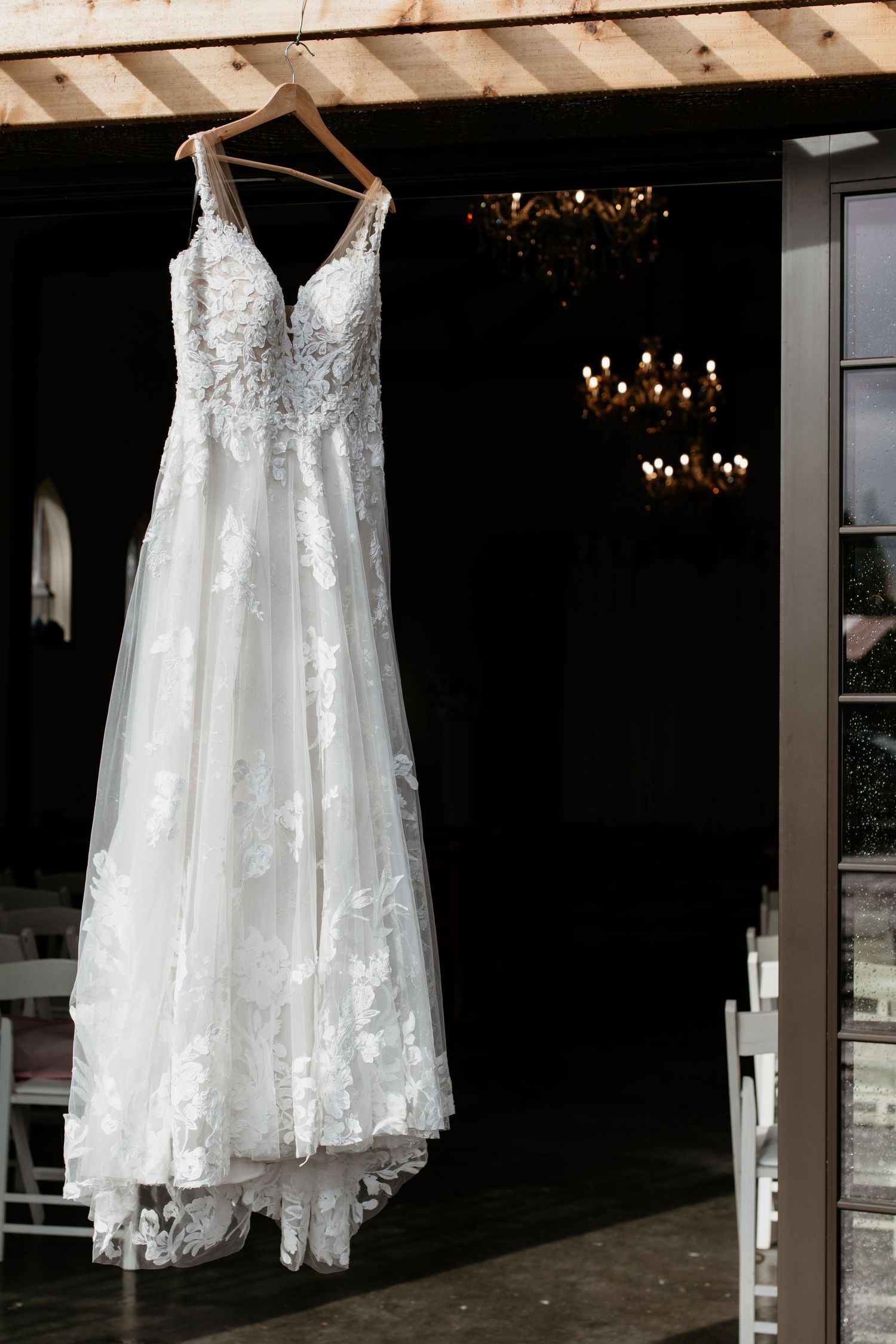 Elegant lace A-line wedding dress hanging on wooden hanger in front of chandelier lit wedding venue.
