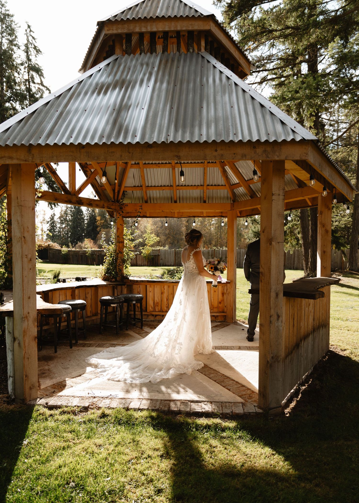 Bride in lace gown stands inside rustic wooden gazebo with corrugated metal roof at outdoor wedding venue.