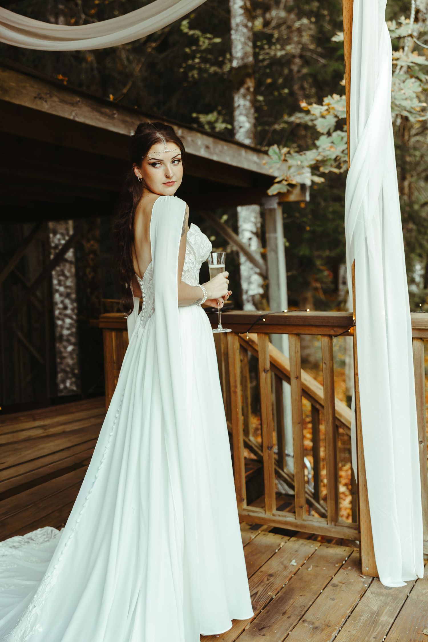 Bride in a flowing white gown with long cape sleeves holds a champagne flute on a rustic wooden deck surrounded by forest.
