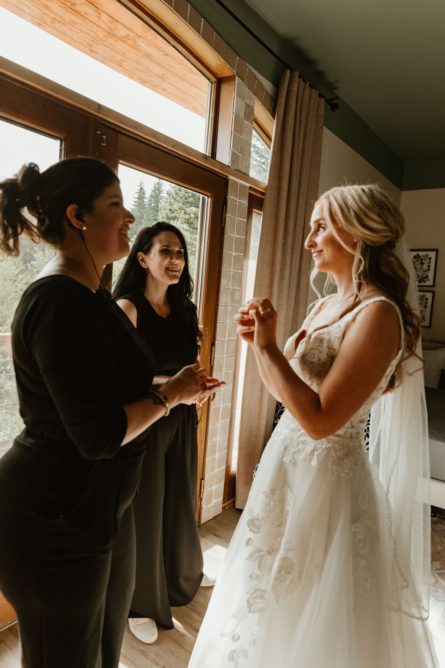 Bride in white lace gown smiling with two women in black during wedding preparations indoors.