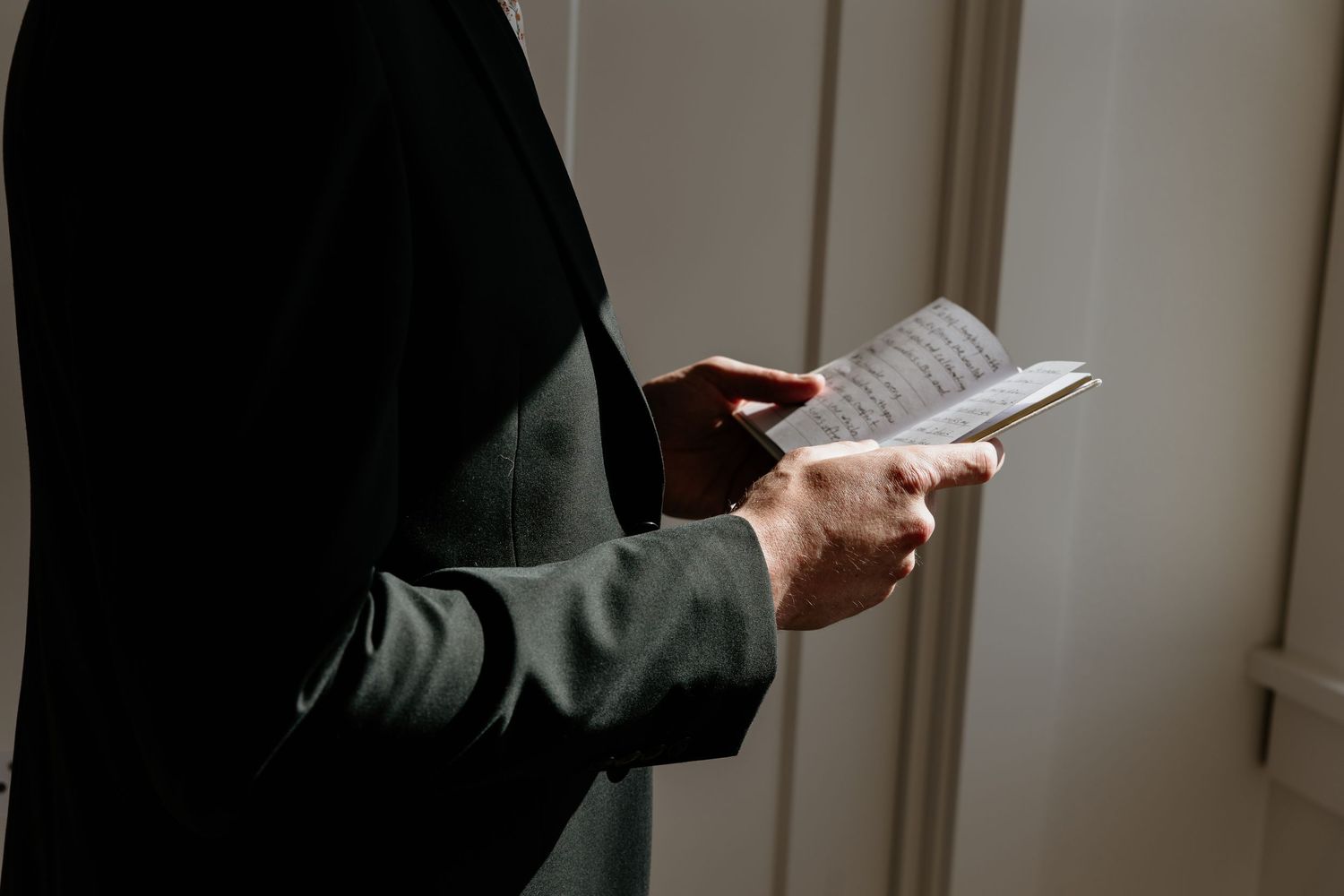 Person in dark suit reading a small book or notepad, holding it with both hands in soft natural light.