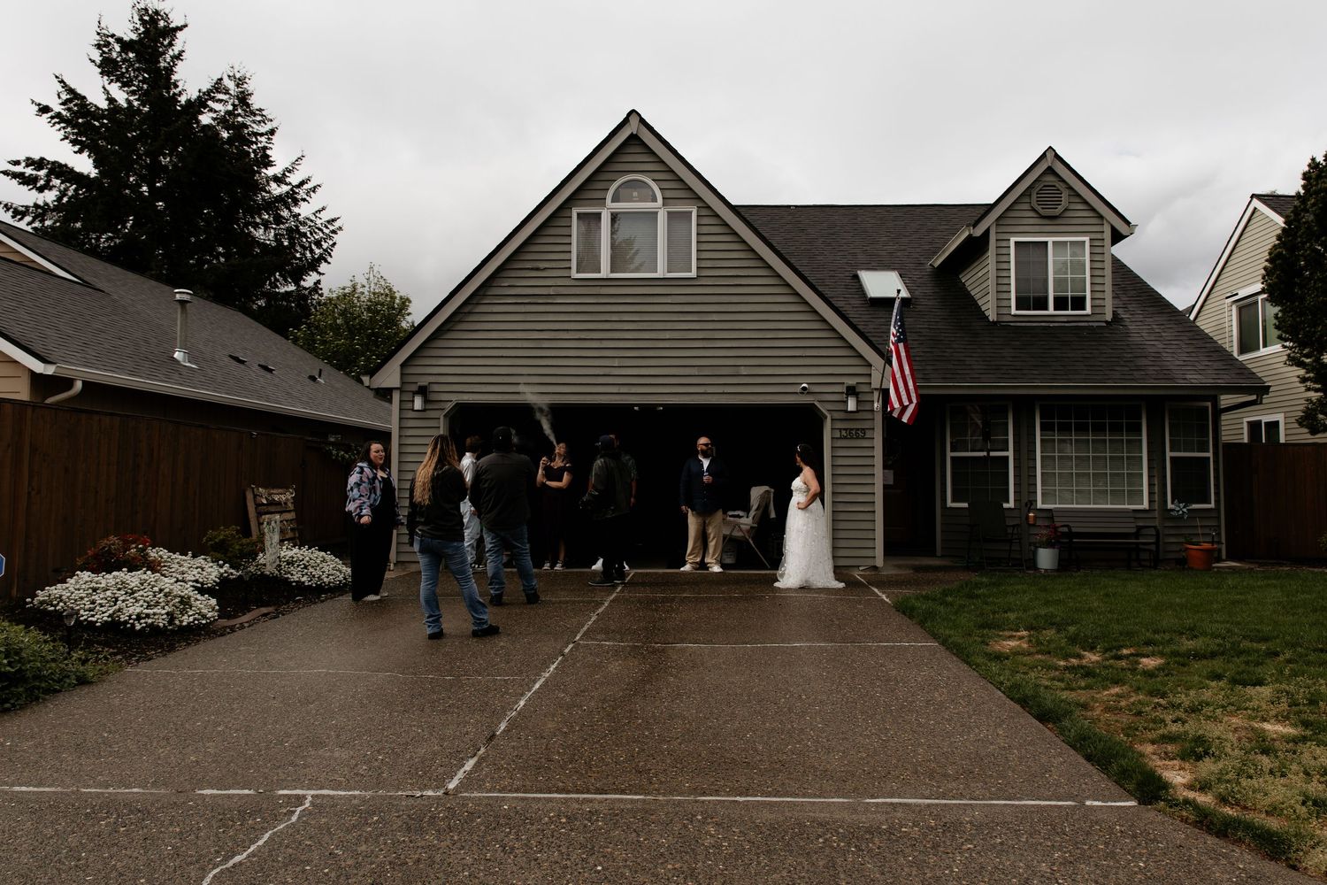 Wedding couple poses in front of a suburban home with an American flag, surrounded by guests on a cloudy day.