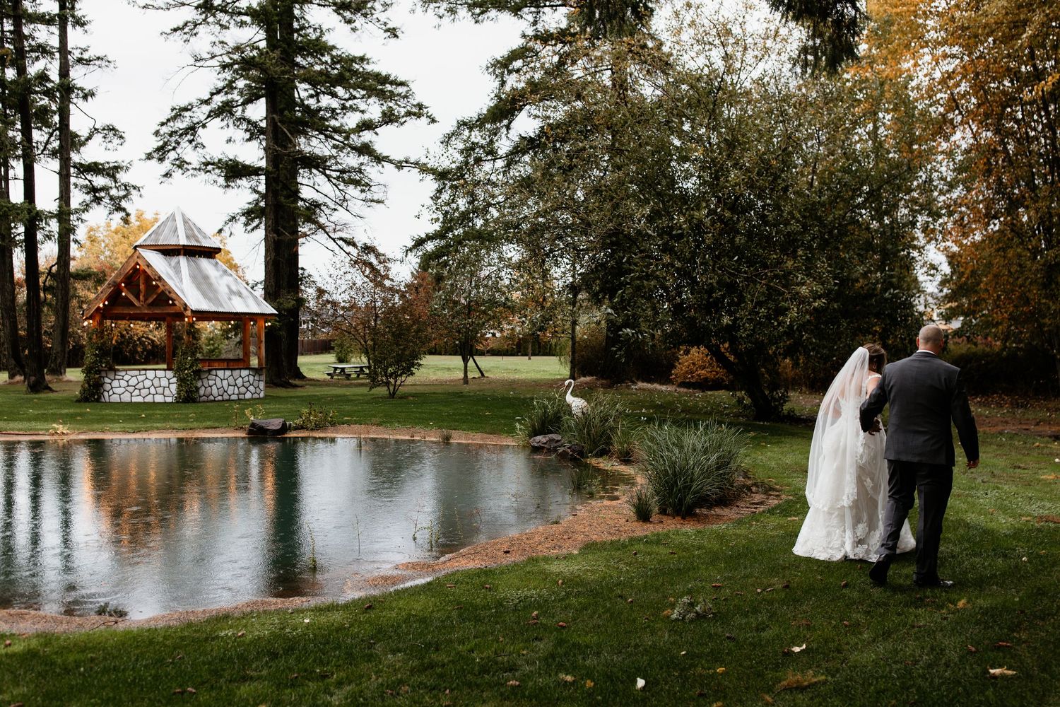 Bride and groom stroll toward a charming gazebo beside a pond.