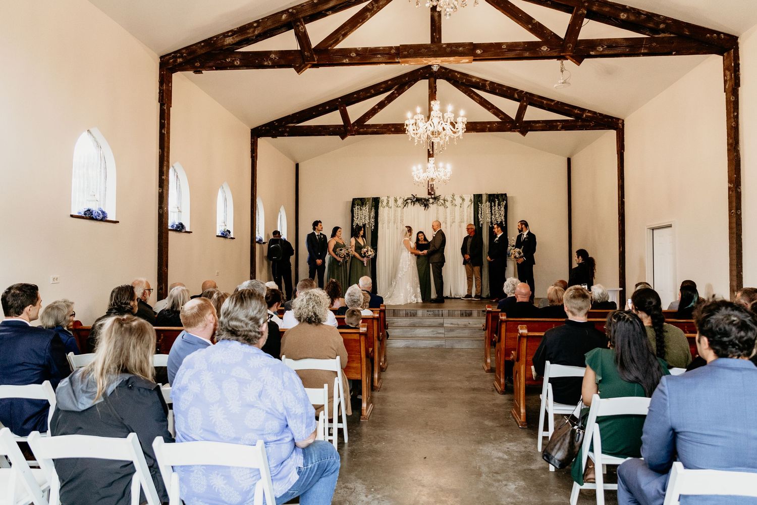 Wide-angle view of wedding ceremony in a white chapel with wood beams, chandelier, and seated guests.