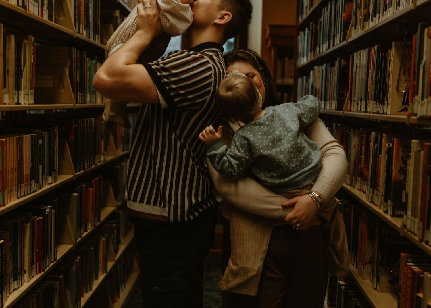 A family sharing a tender moment between library bookshelves with a parent holding a young child.