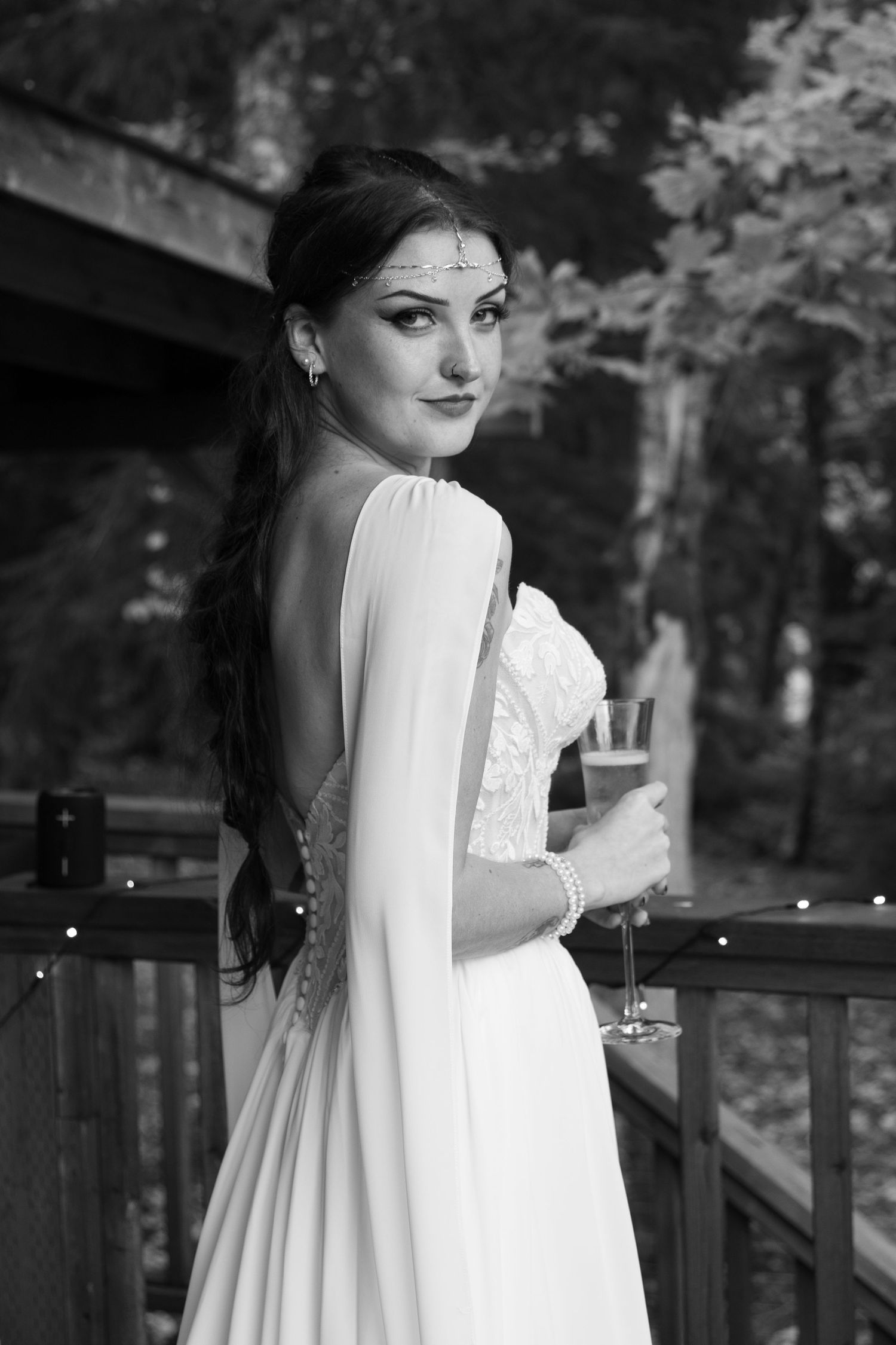 Black and white portrait of a bride in a lace gown with an open back and cape sleeves, holding a champagne glass on a wooden deck.
