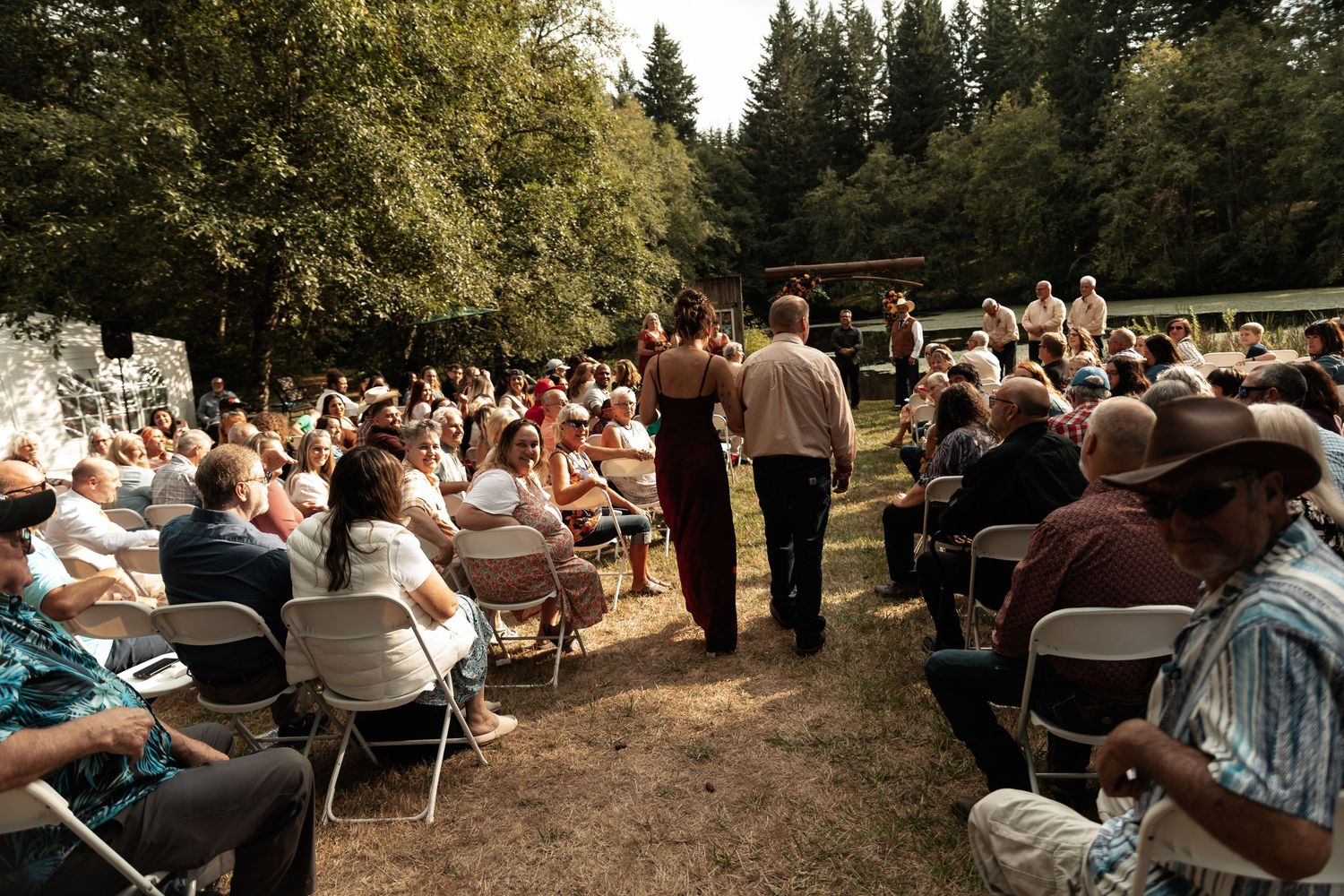 Large outdoor audience seated on chairs at a shaded woodland gathering or performance event on a sunny day.