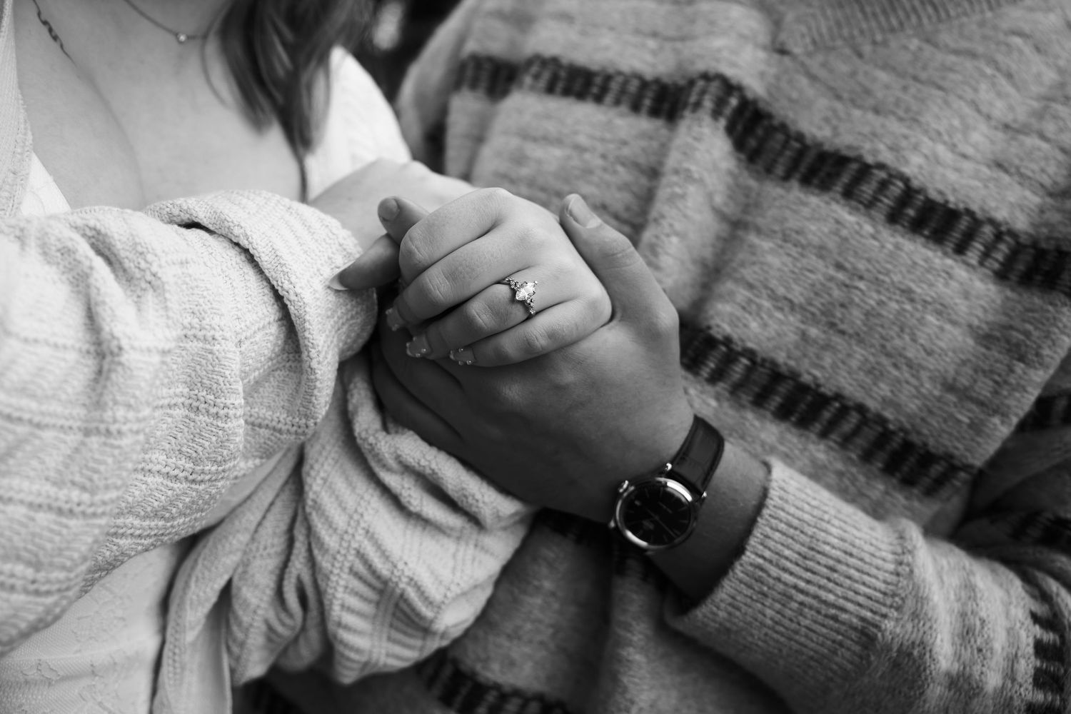 Black and white close-up of a couple holding hands, her engagement ring and his wristwatch visible, both in cozy knit sweaters.