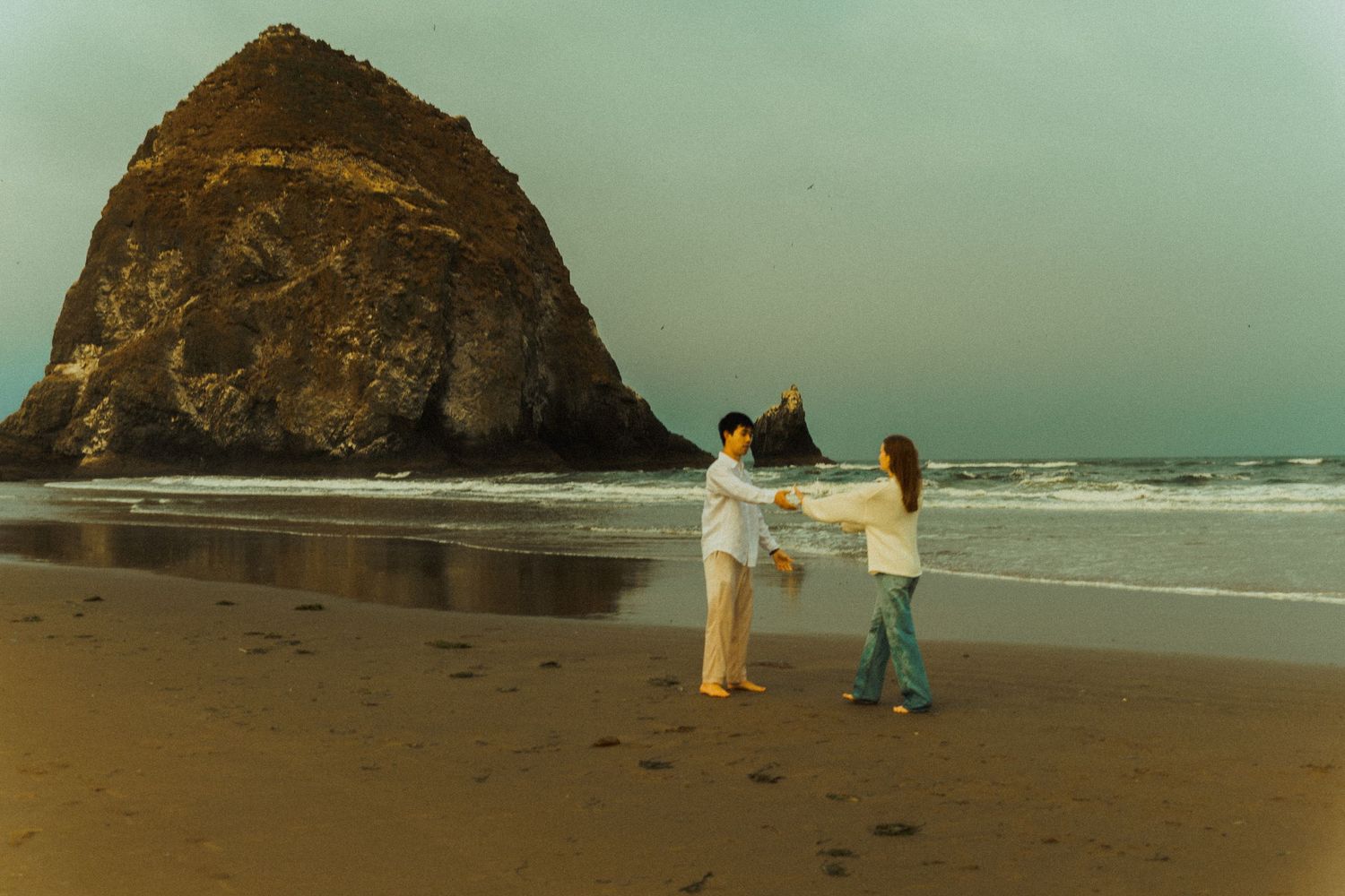 Two people hold hands and dance on a wet sandy beach at dusk, with a large sea stack rising from the shoreline and gentle waves behind them.