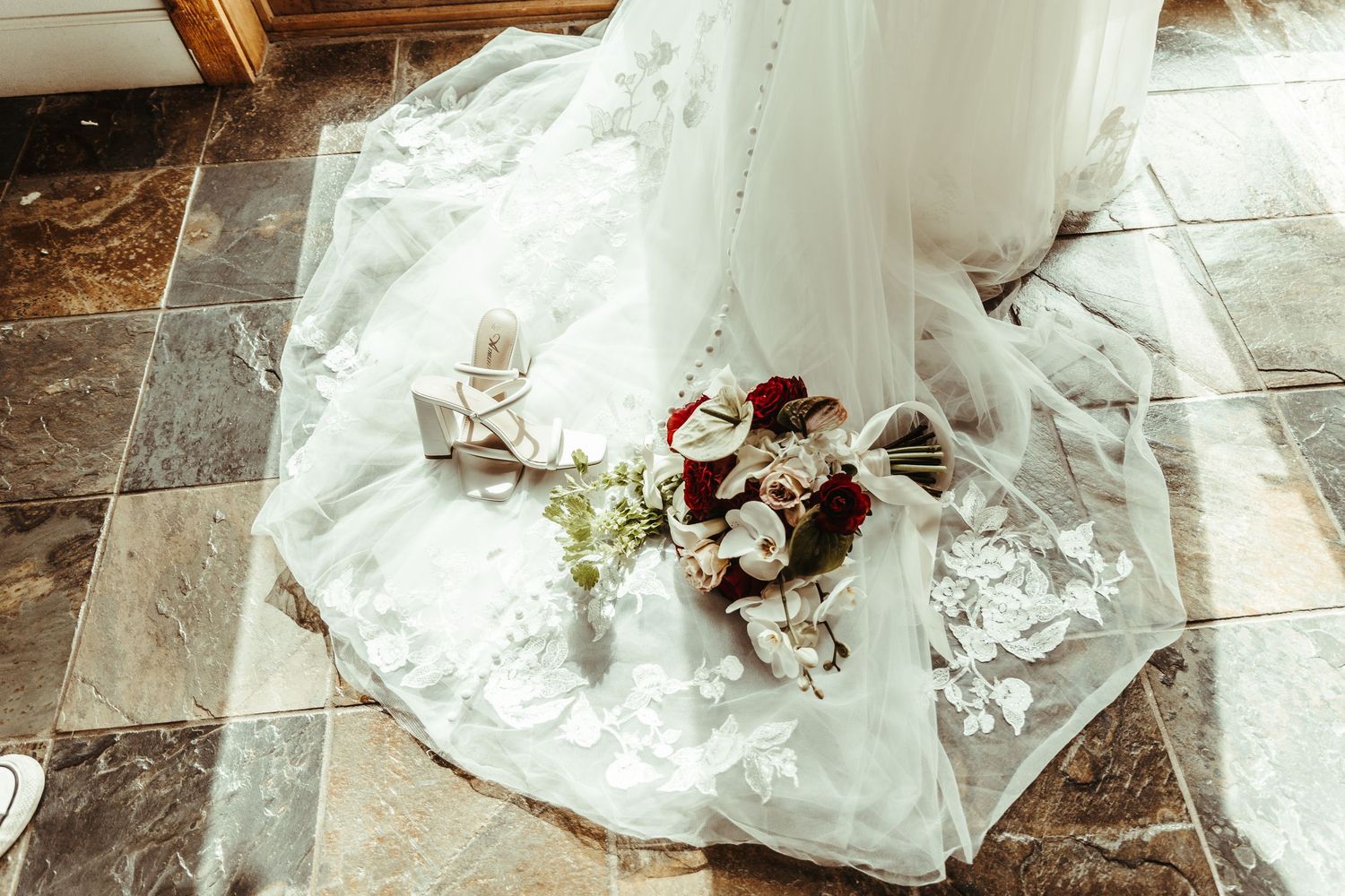 Elegant wedding dress train spread on stone floor with bridal bouquet of red and white flowers and heels.