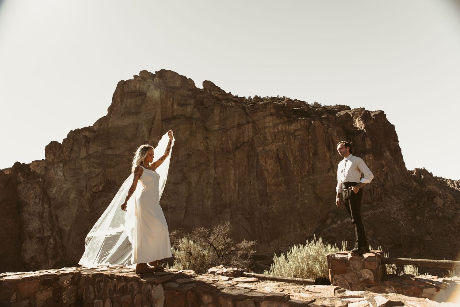 Bride in white gown and groom in desert canyon wedding photo with dramatic red rock formations backdrop.